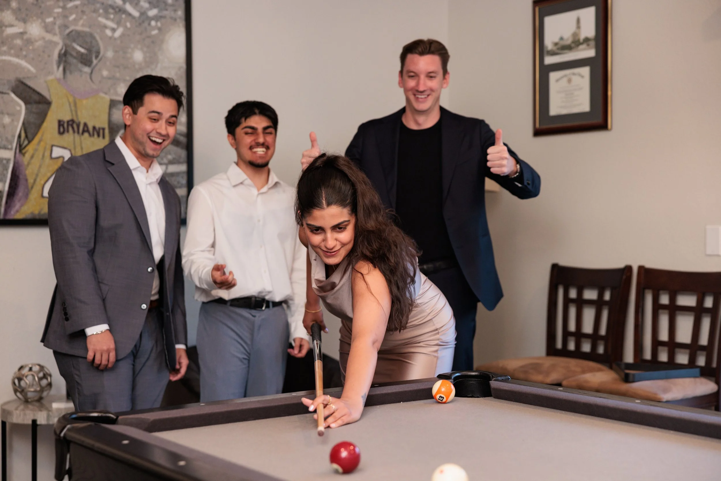 A woman is playing billiards while four men in formal attire watch and smile, with one giving a thumbs-up in the background.