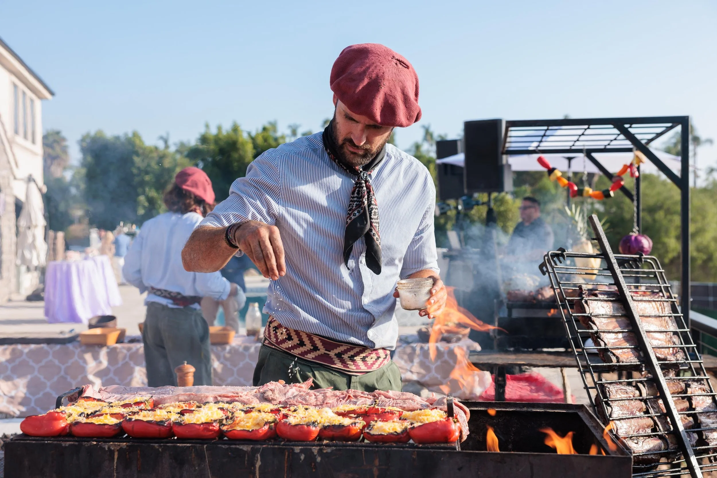 Man grilling stuffed tomatoes and meat at outdoor barbecue.