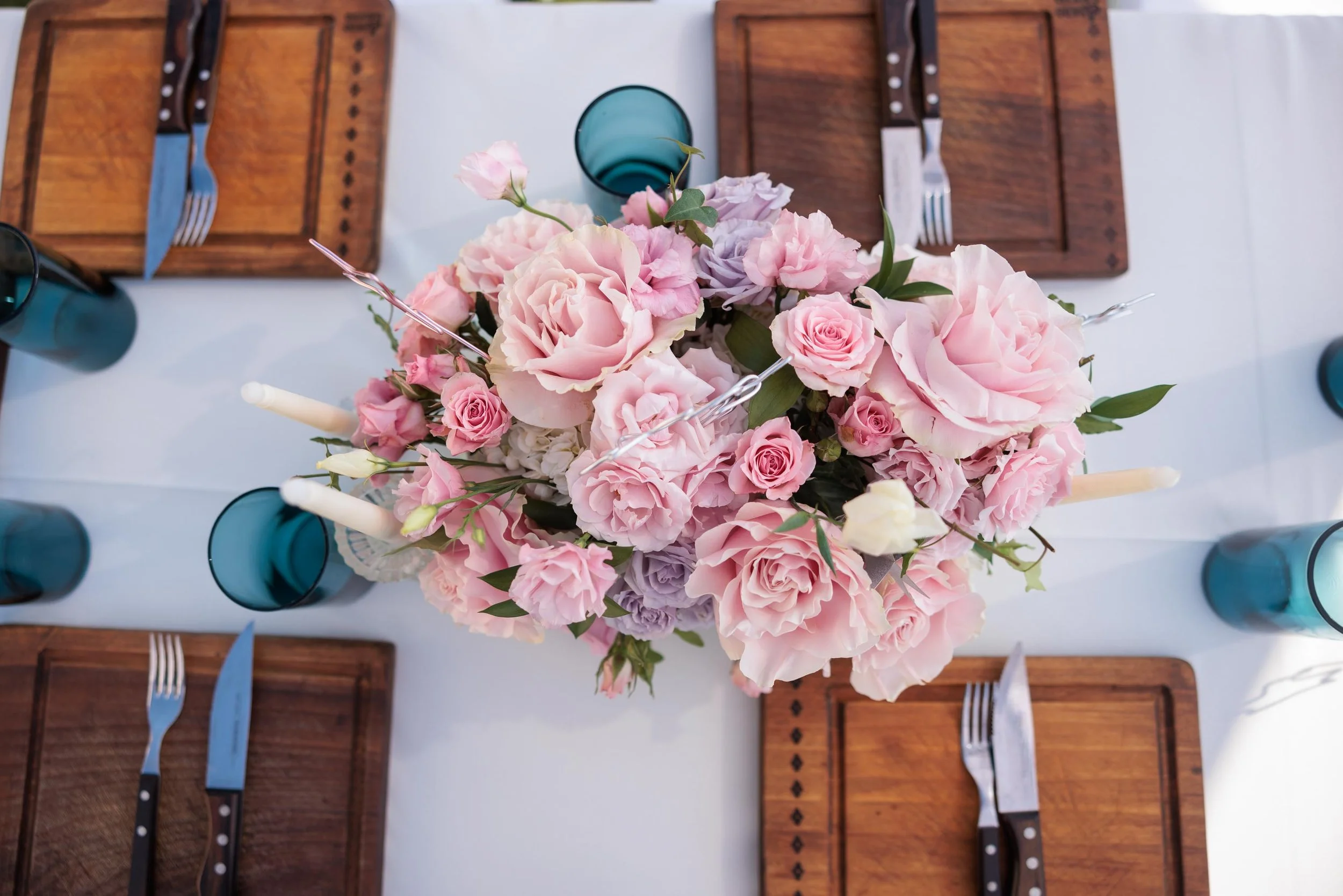 A top view of a dining table with a floral centerpiece of pink and purple roses and peonies, surrounded by wooden placemats, blue glasses, white candles, and silverware with wooden handles.