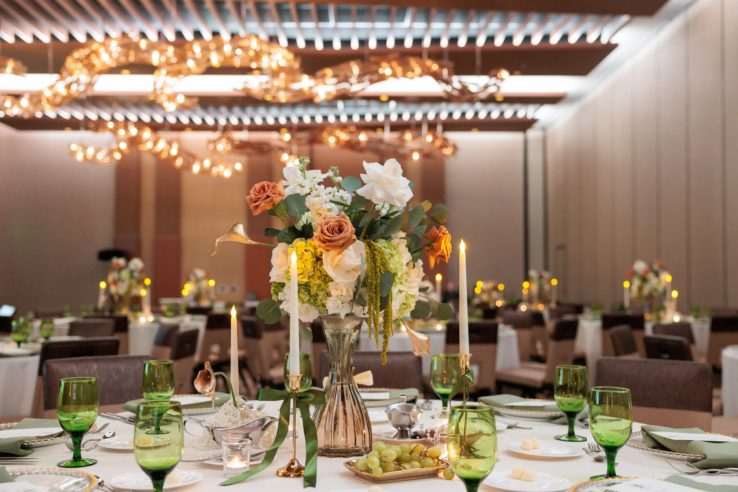 Elegant banquet table decorated with a large flower arrangement in a glass vase, surrounded by lit white candles, green wine glasses, and small plates of grapes, set in a decorated reception hall with warm lighting and ceiling drapes.