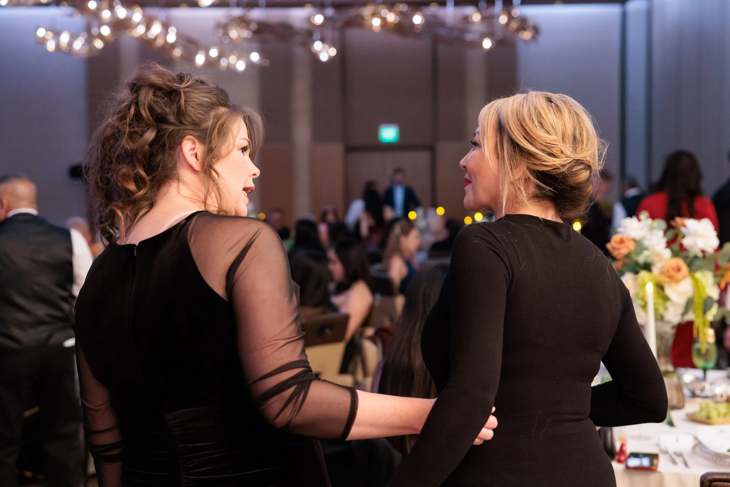 Two women are talking and holding hands at a formal event or celebration in a banquet hall.