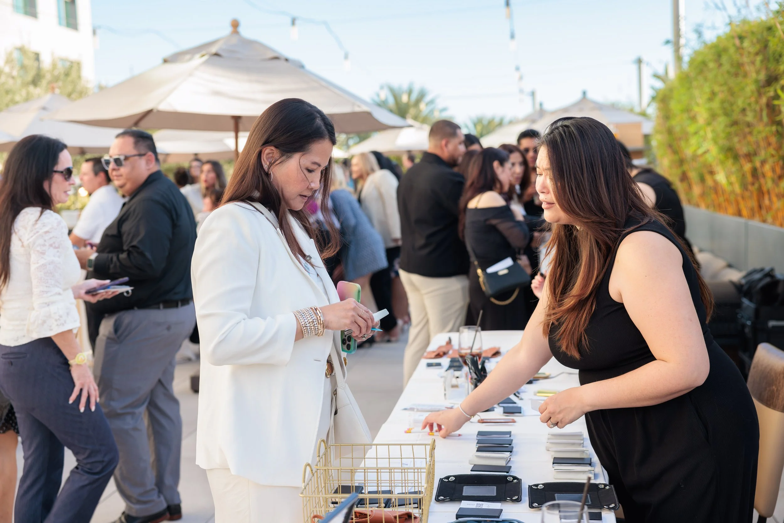 People shopping at an outdoor market table with accessories, with other shoppers and umbrellas in the background.