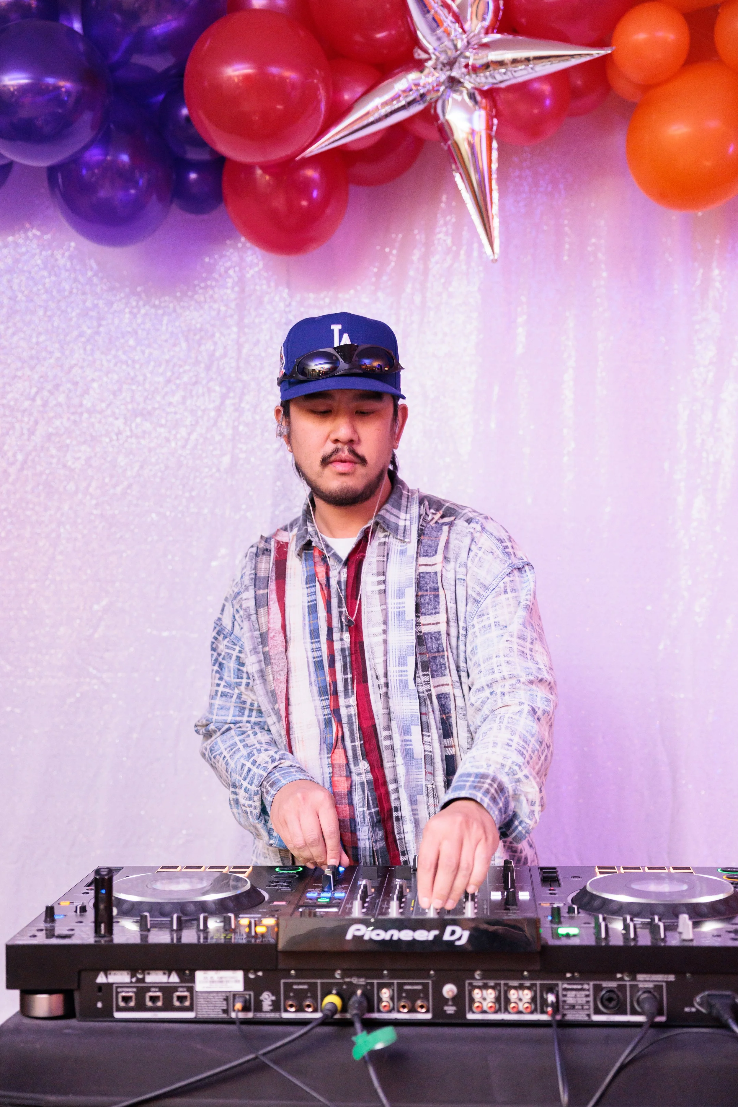 A DJ wearing a baseball cap and sunglasses, DJing at a party with purple and orange balloons and star-shaped decorations in the background.
