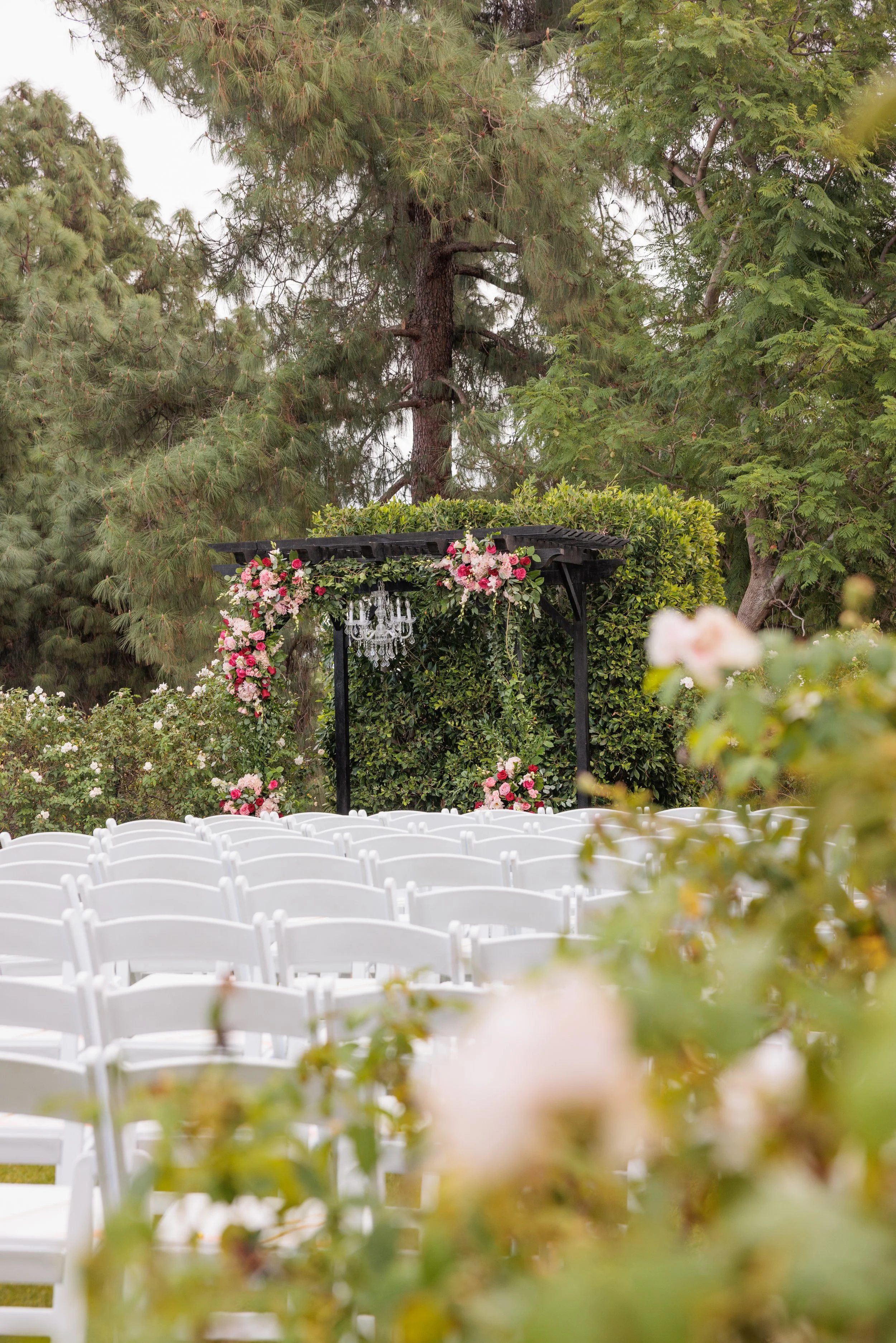 Outdoor wedding ceremony setup with white chairs, floral arch with pink and white flowers, and chandeliers hanging from a black canopy, surrounded by green trees and bushes.