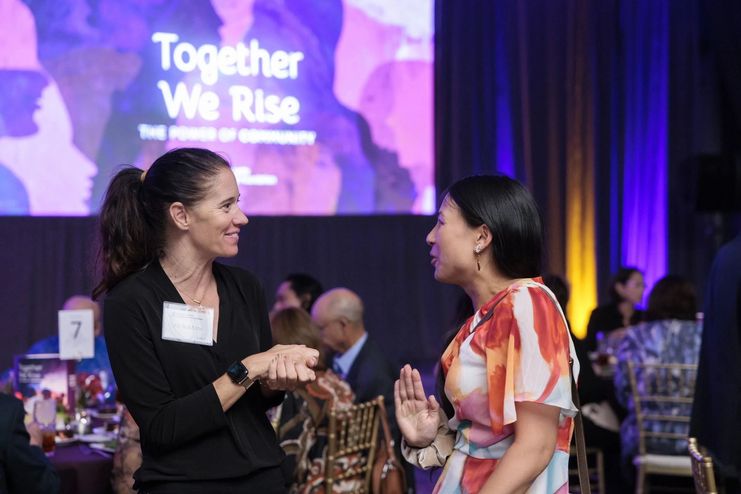 Two women engaged in conversation at a formal event with a large screen in the background displaying the text "Together We Rise," with additional text that is partially obscured.