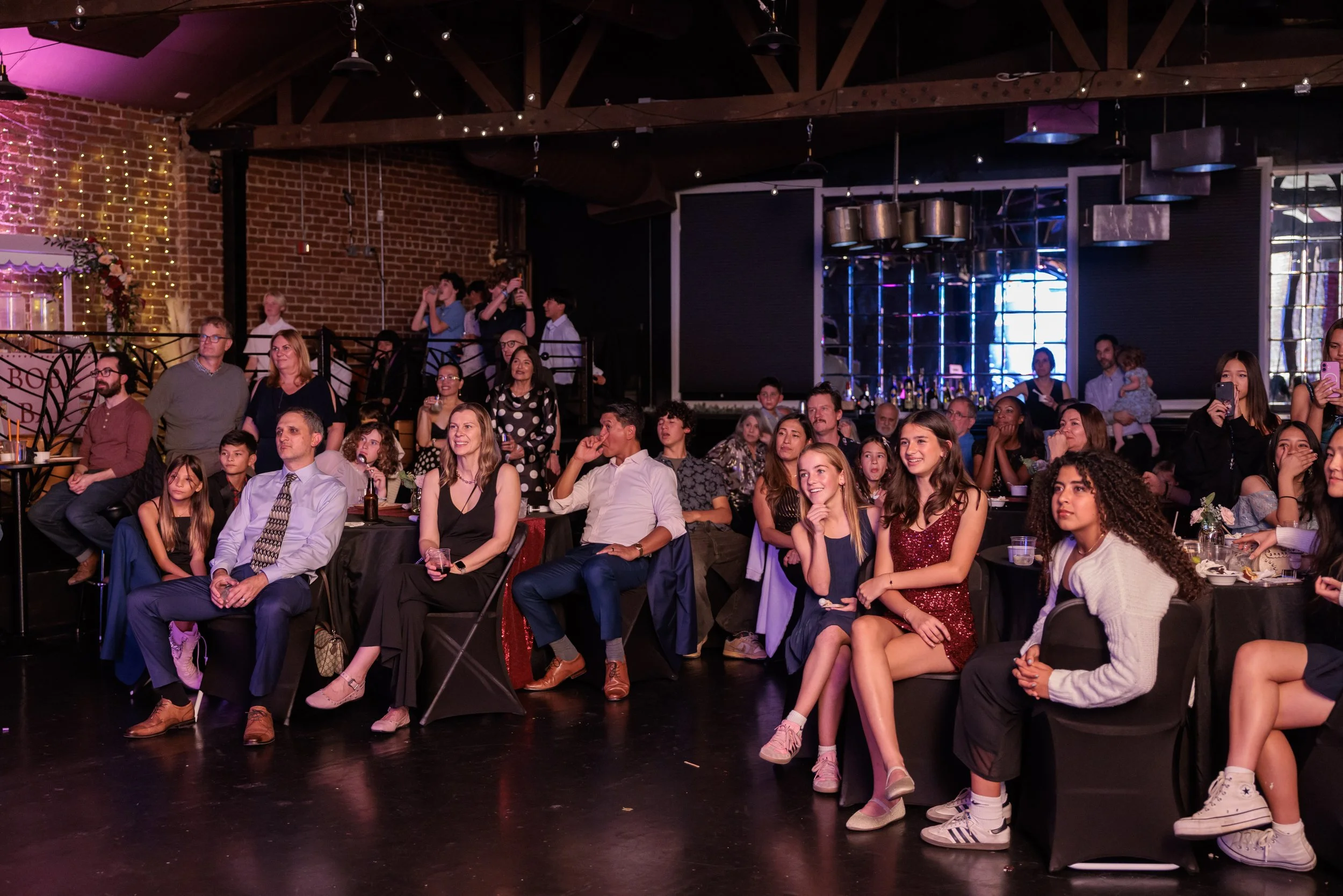 Audience seated at tables and standing, watching a performance in a dimly lit venue with brick walls and string lights.