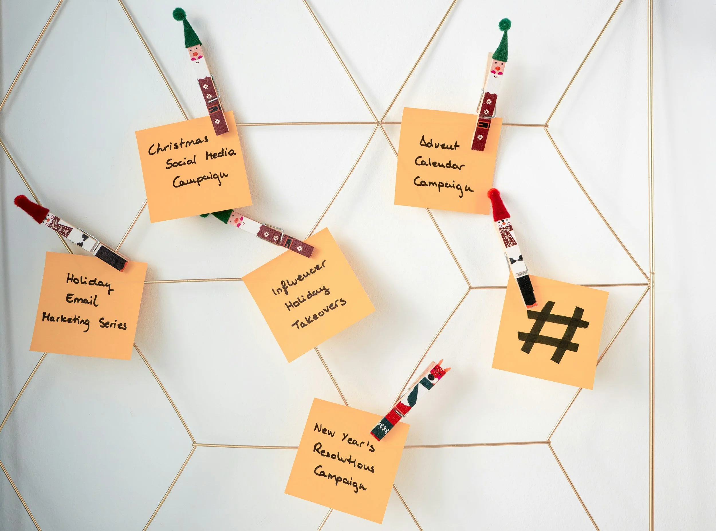 A white wire grid decorated with orange sticky notes held by clothespins with festive designs, featuring handwritten labels for holiday-themed social media campaigns and planning.