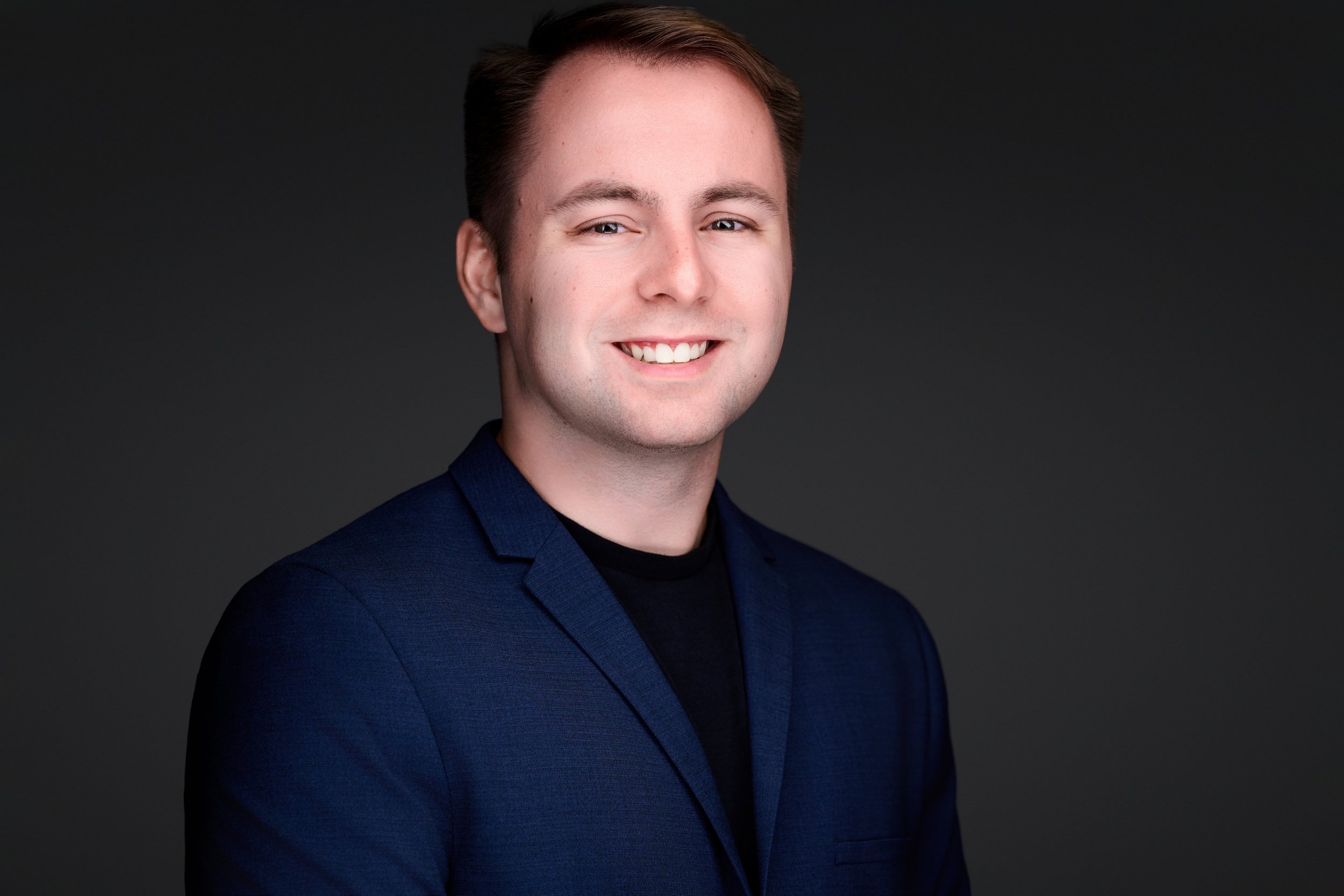 Professional headshot of a young man with short brown hair, wearing a dark blue blazer and black shirt, smiling against a dark background.