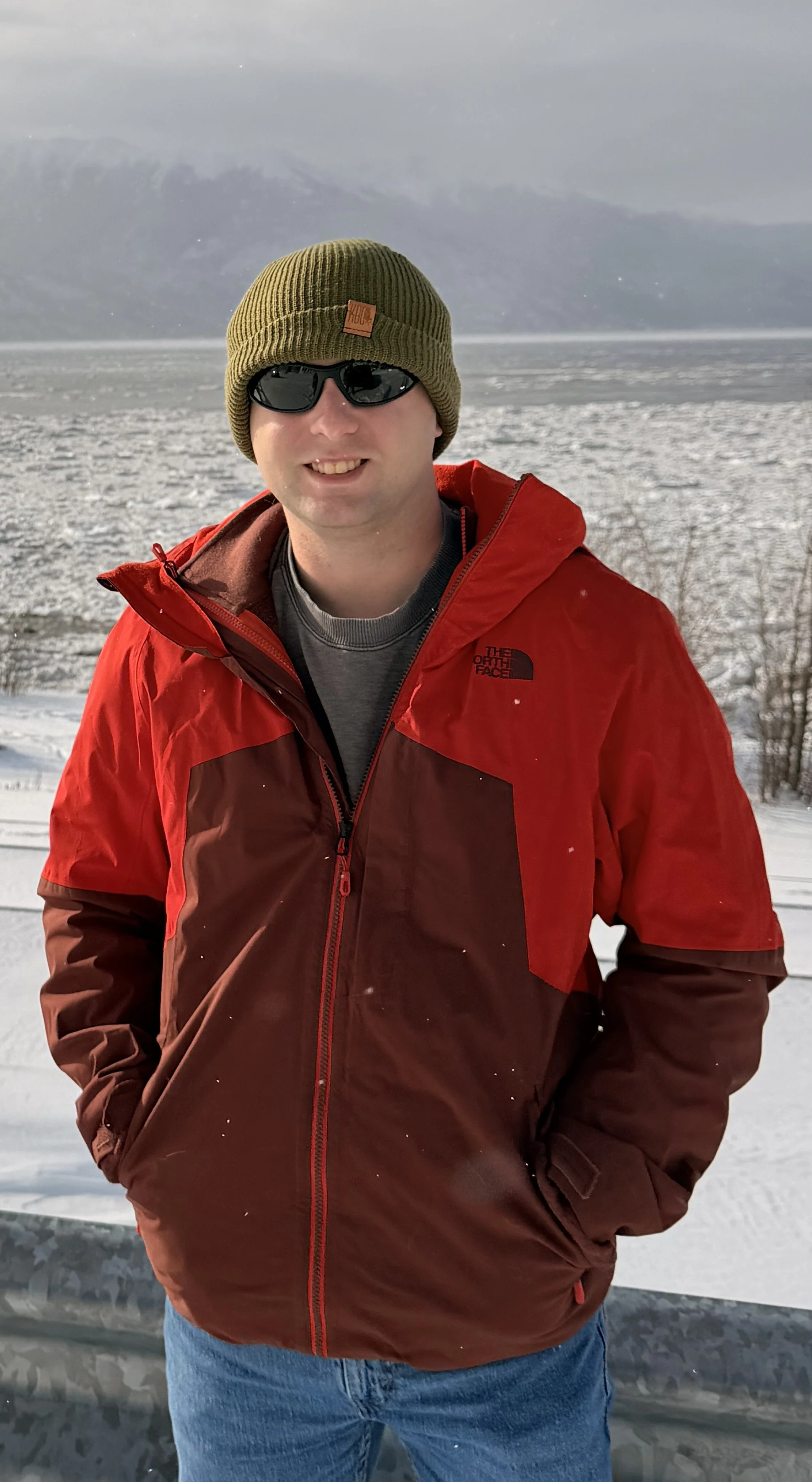 Man in a red and brown jacket, olive green beanie, and sunglasses standing by a snowy landscape with mountains in the background.