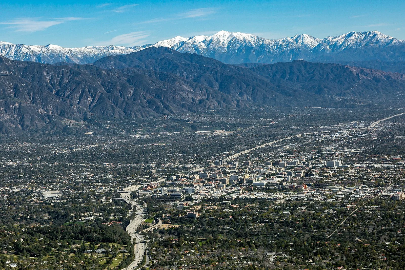 Aerial view of a city with highways and buildings, mountains in the background, some snow-capped peaks.