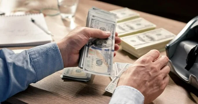 Person counting stacks of hundred-dollar bills on a wooden table with more cash and a money counting machine nearby.