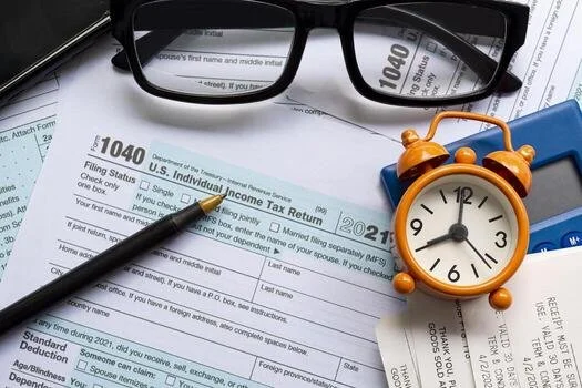 A pair of black eyeglasses, an orange alarm clock, and a black pen resting on a U.S. 1040 income tax form with documents and papers underneath.