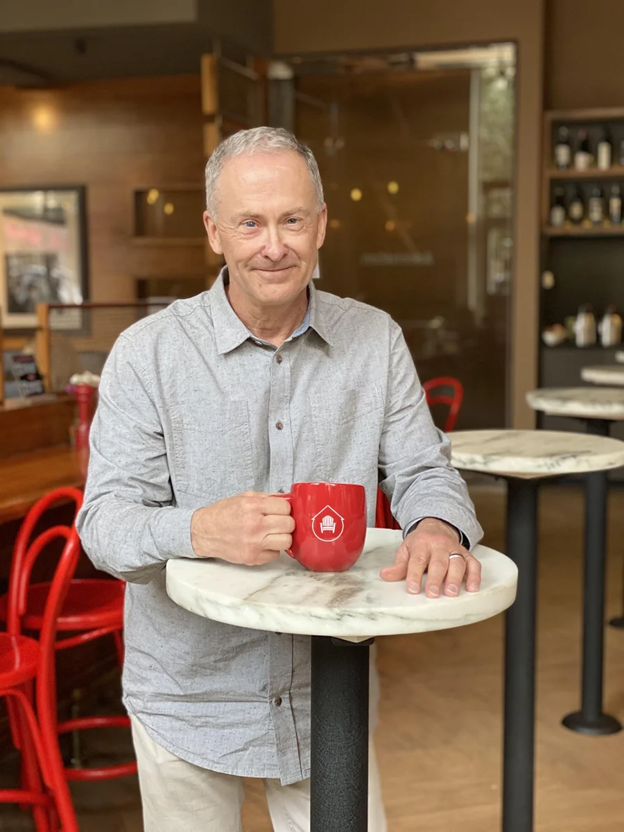 The Kahler Team real estate team leader and Keller Williams Black Hills owner, David Kahler, wearing a light gray button-up shirt standing at a marble-topped table in a cozy cafe, holding a red mug with the teams icon on it, smiling at the camera.