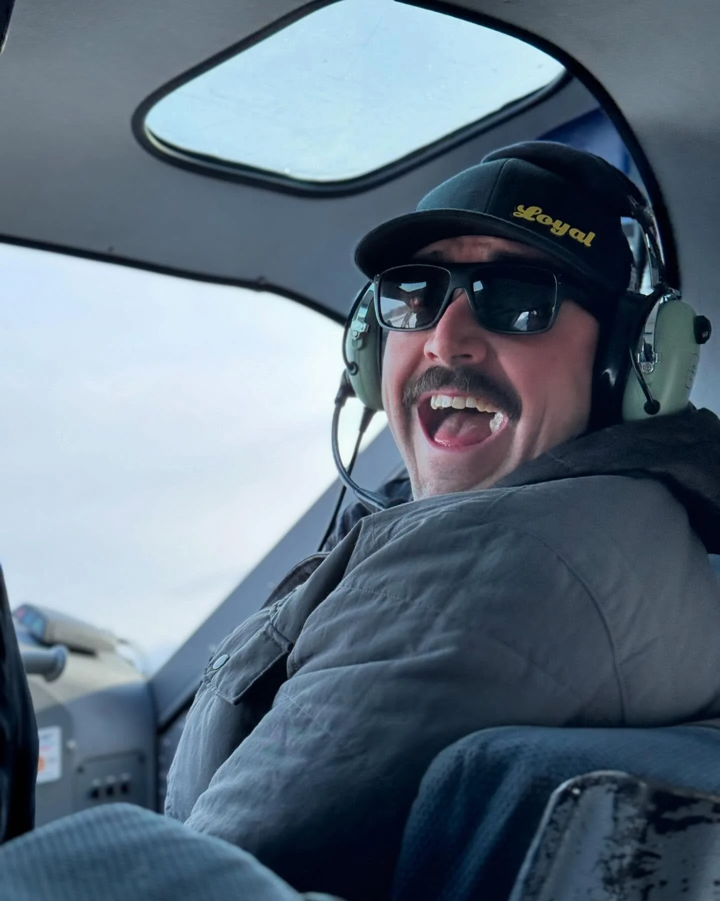 Loyal founder Mark Falcon, sporting a mustache and loving his brand wearing a baseball cap with 'Loyal' written on it, sunglasses, and a headset, smiling and sitting inside a helicopter.
