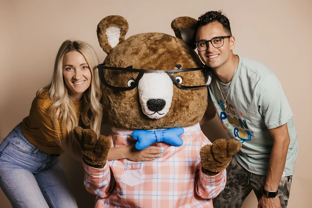 Nerdy Nuts Peanut Butter Co-Founders, Craig and Ericka Mount, posing with Benny the Bear maskot, wearing a pink plaid shirt, and a blue bow tie against a beige background.