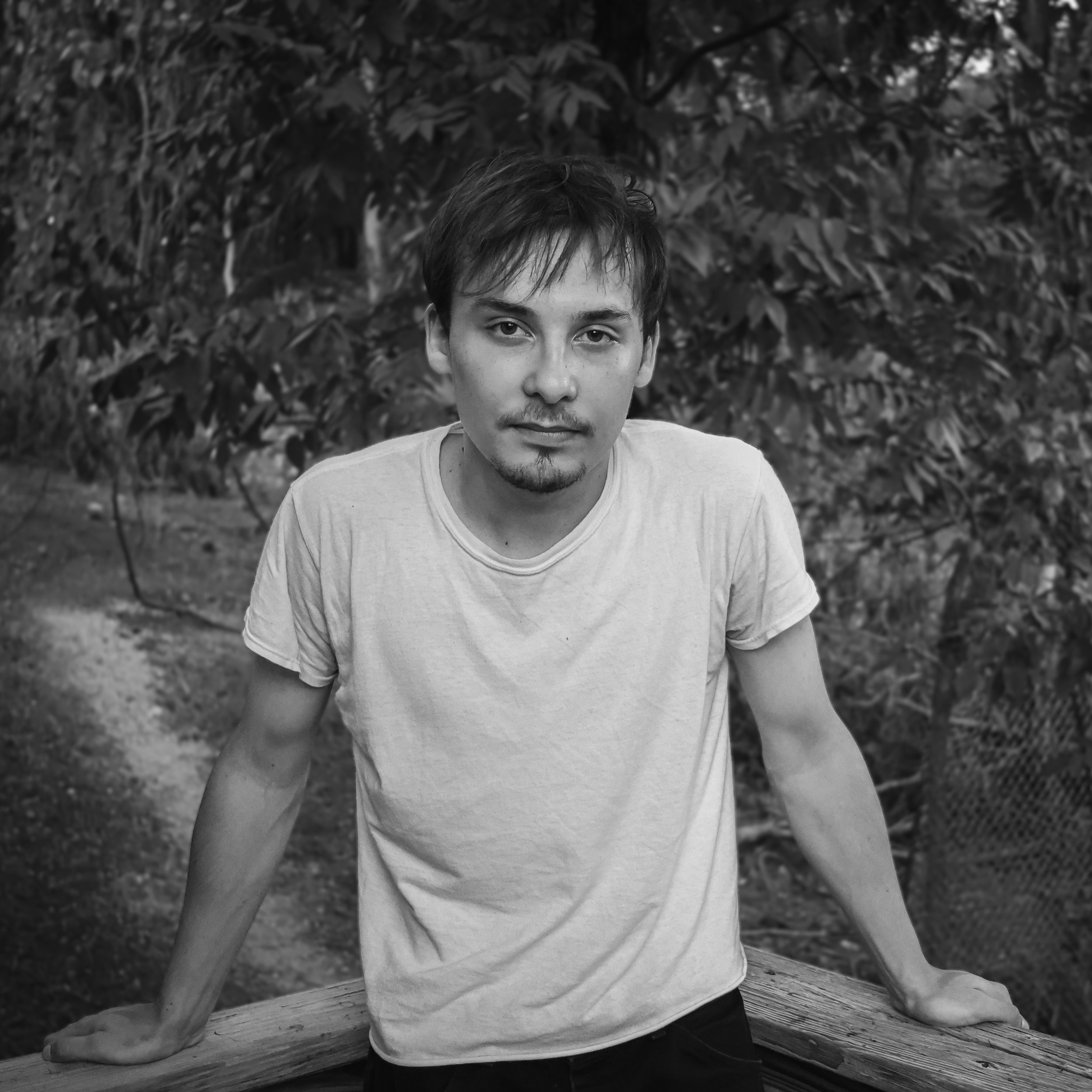 A young man with short hair and a slight goatee posing outdoors, leaning on a wooden railing with trees in the background.