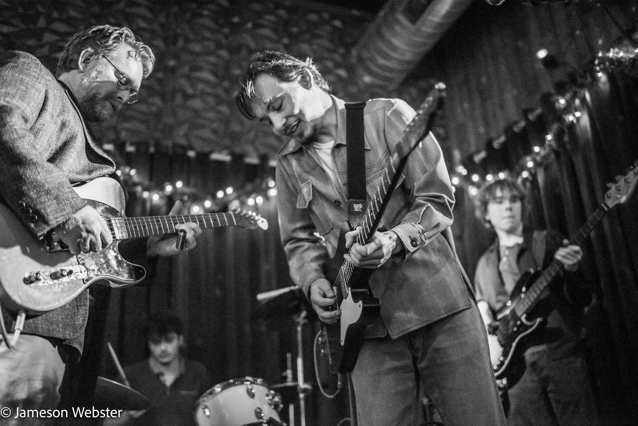 Three musicians playing electric guitars and drums in a performance with string lights and curtains in the background.