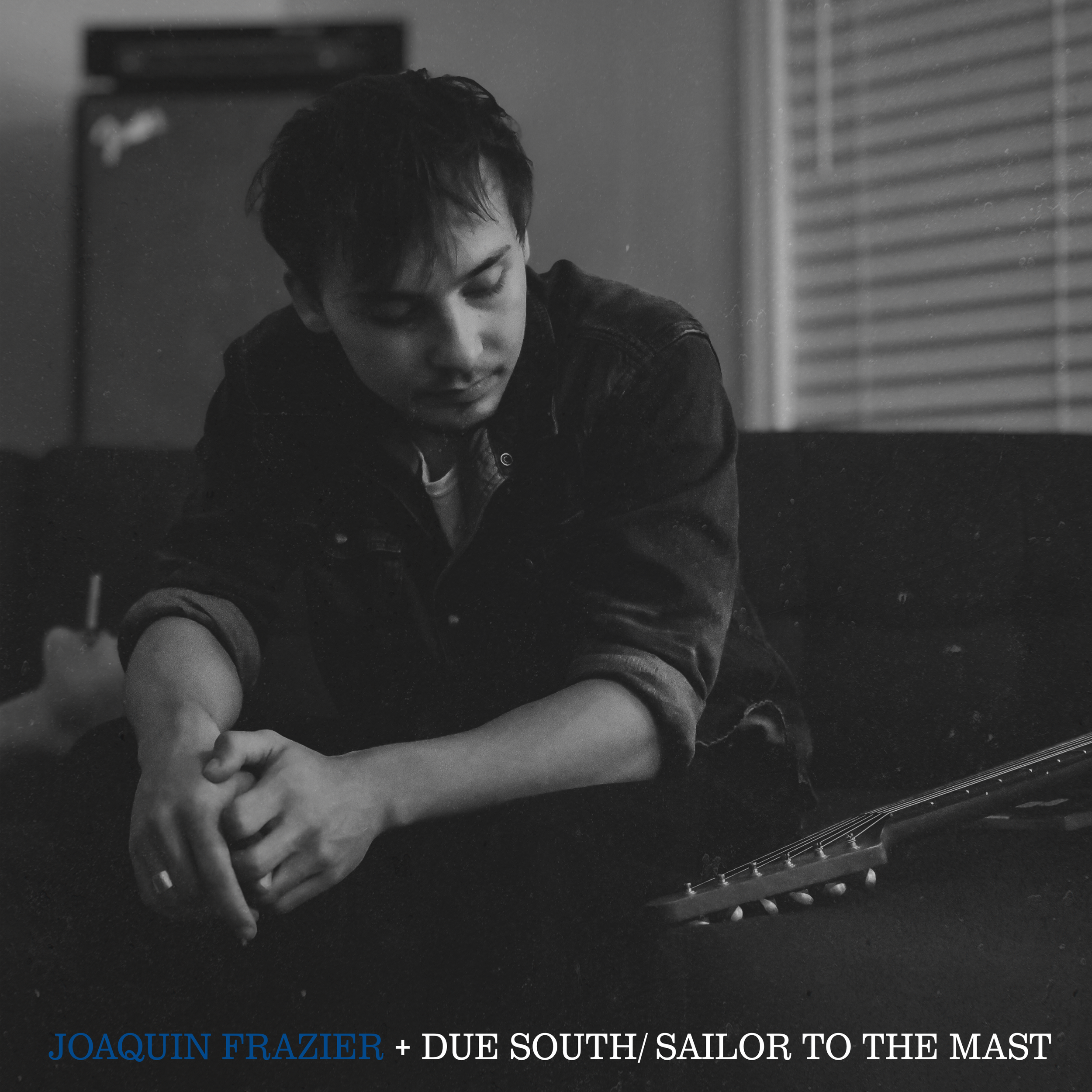 Black and white photograph of a young man with short dark hair, wearing a denim jacket, sitting with a guitar on his lap, looking down thoughtfully in a dimly lit room with blinds in the background.