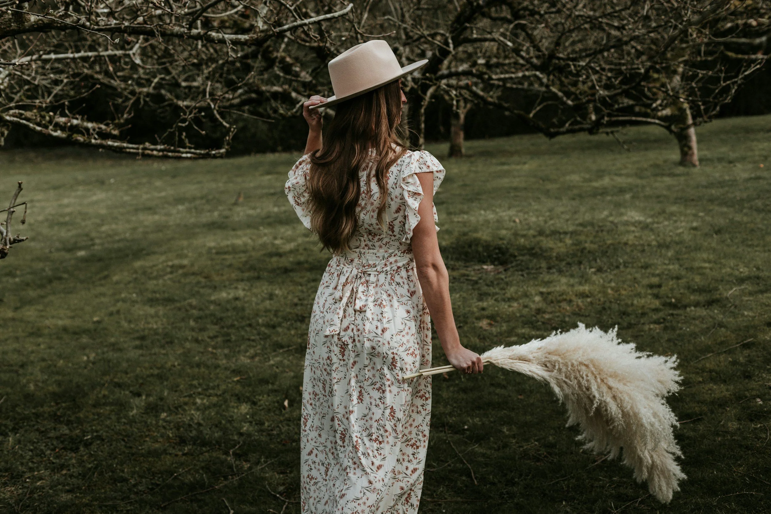 A woman with long wavy hair, wearing a floral dress and a wide-brimmed hat, holding a feather duster, standing outdoors in a grassy area surrounded by trees.
