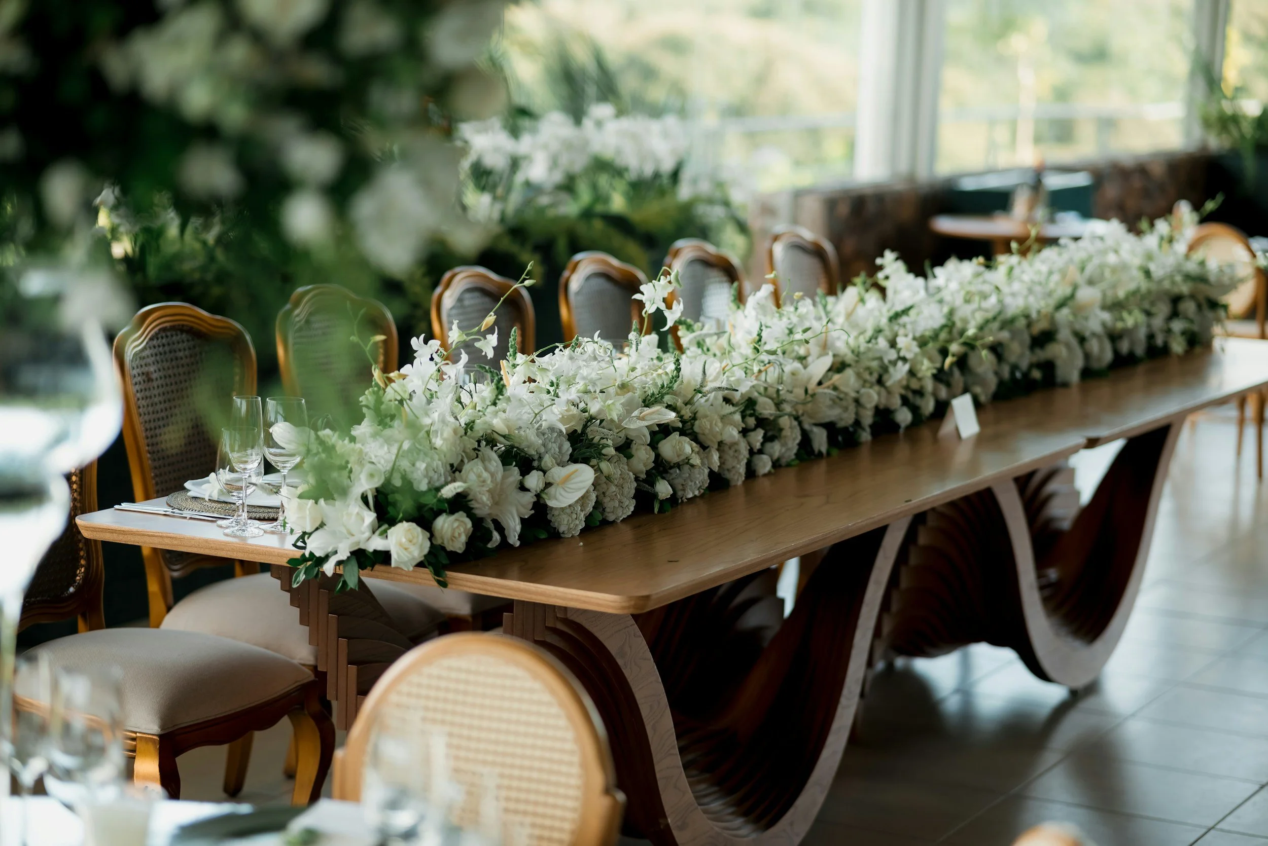 A long wooden dining table decorated with a lush white flower arrangement, set with glassware and plates, in a bright room with large windows and green scenery outside.