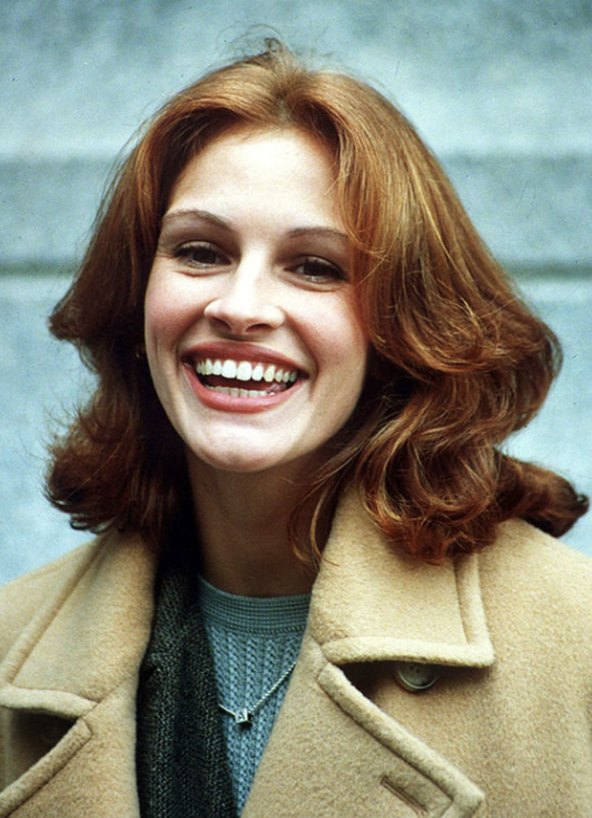 Close-up of a woman with red hair smiling, wearing a beige coat and a gray sweater.
