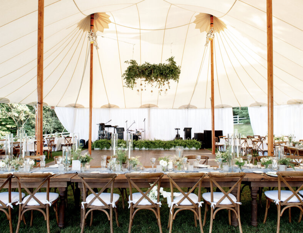 Wedding reception setup inside an ivory sailcloth tent with wooden chairs and a decorated table, with a stage and musical instruments in the background.