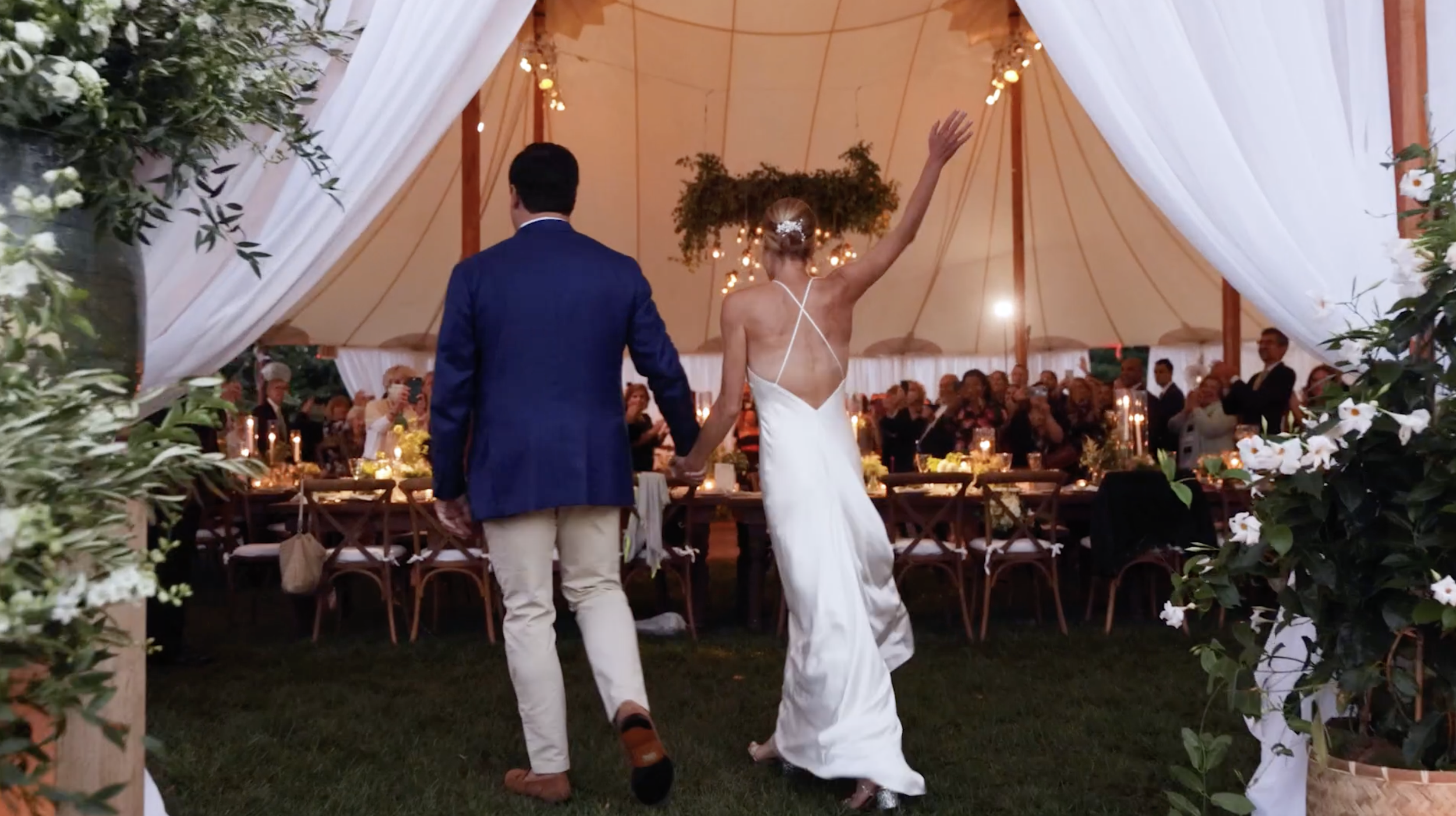 Bride and groom holding hands and walking into a wedding reception tent, bride waving, guests seated at decorated tables.