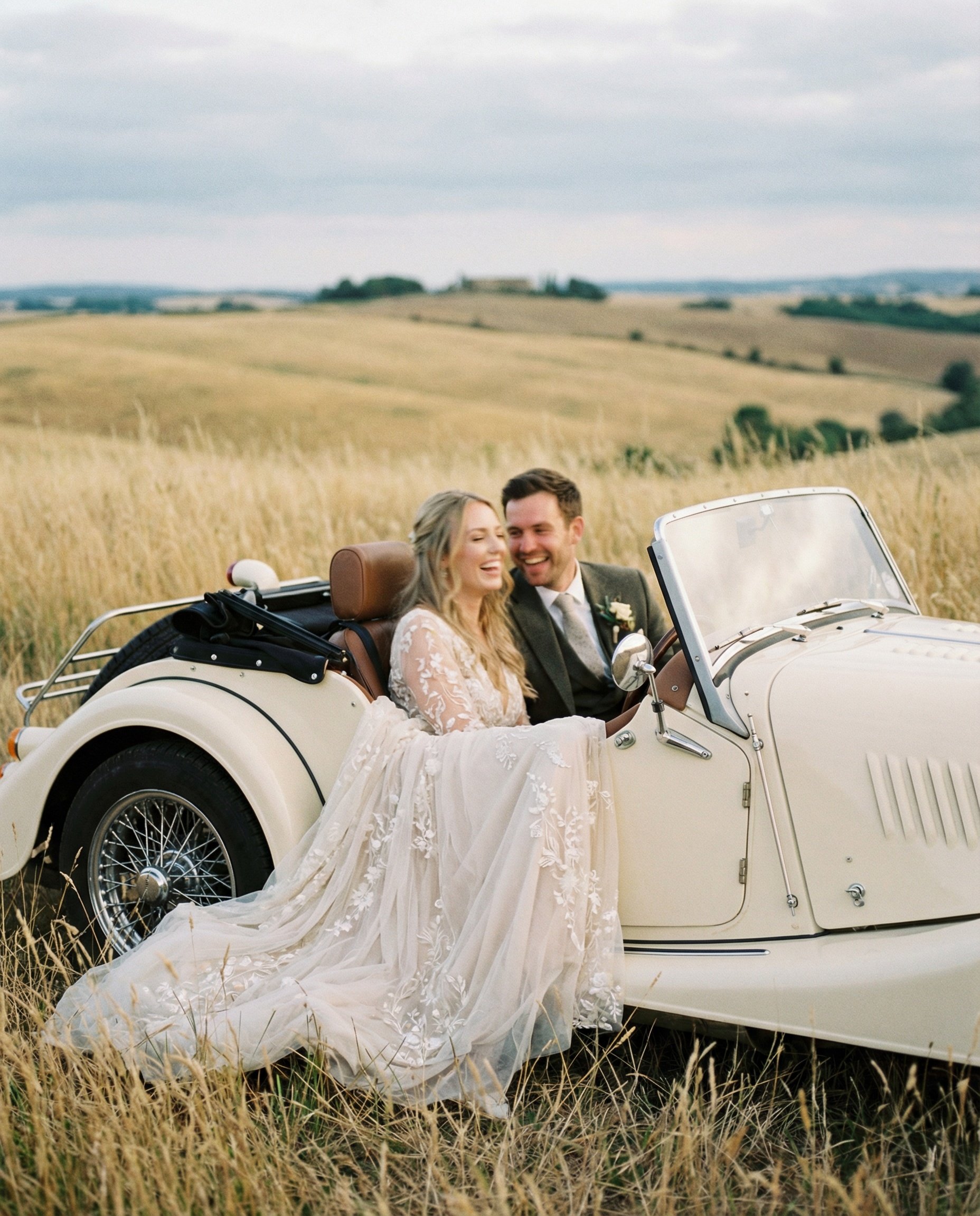 A newlywed couple sitting in a vintage white car in a grassy field, smiling and looking at each other, with rolling hills in the background.