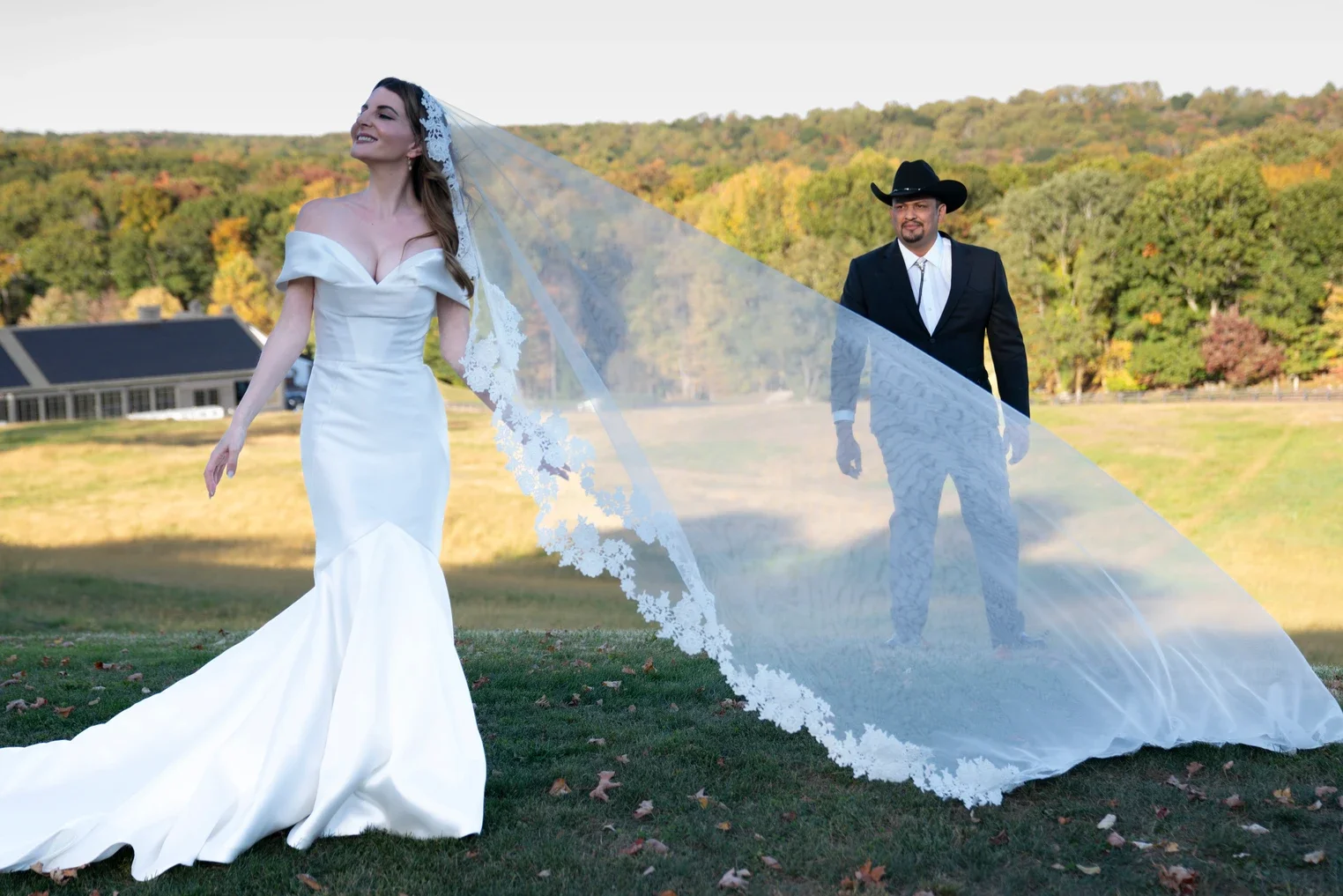 A bride in a white wedding dress walking outdoors with a long train and lace veil, accompanied by a groom in a black suit and cowboy hat, against a backdrop of trees and a house.