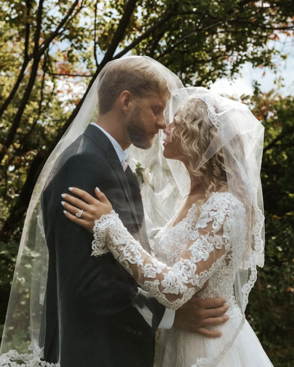 A bride and groom in wedding attire stand close together outdoors, sharing an intimate moment under a veil, with trees and leaves in the background.