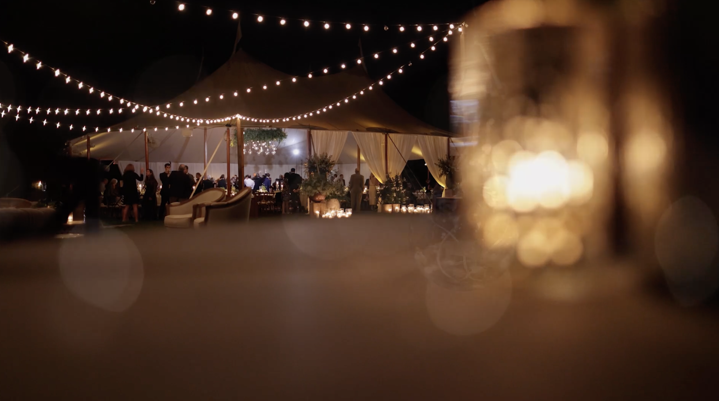 Nighttime outdoor event with string lights overhead, people gathered under a tent with draped curtains, tables, and floral decorations.