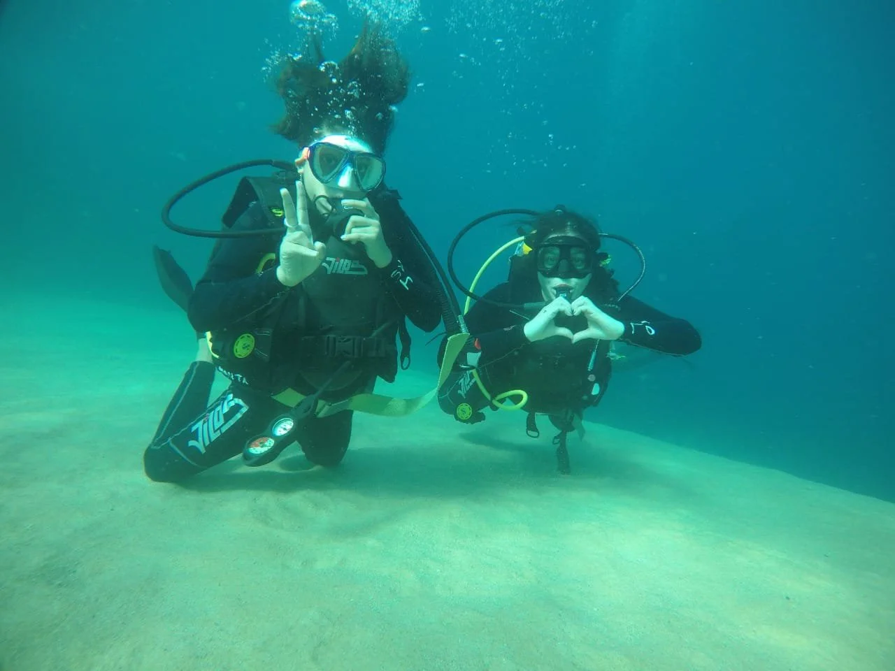Dos personas haciendo buceo en el océano, con equipo de buceo y formando corazones con sus manos.
