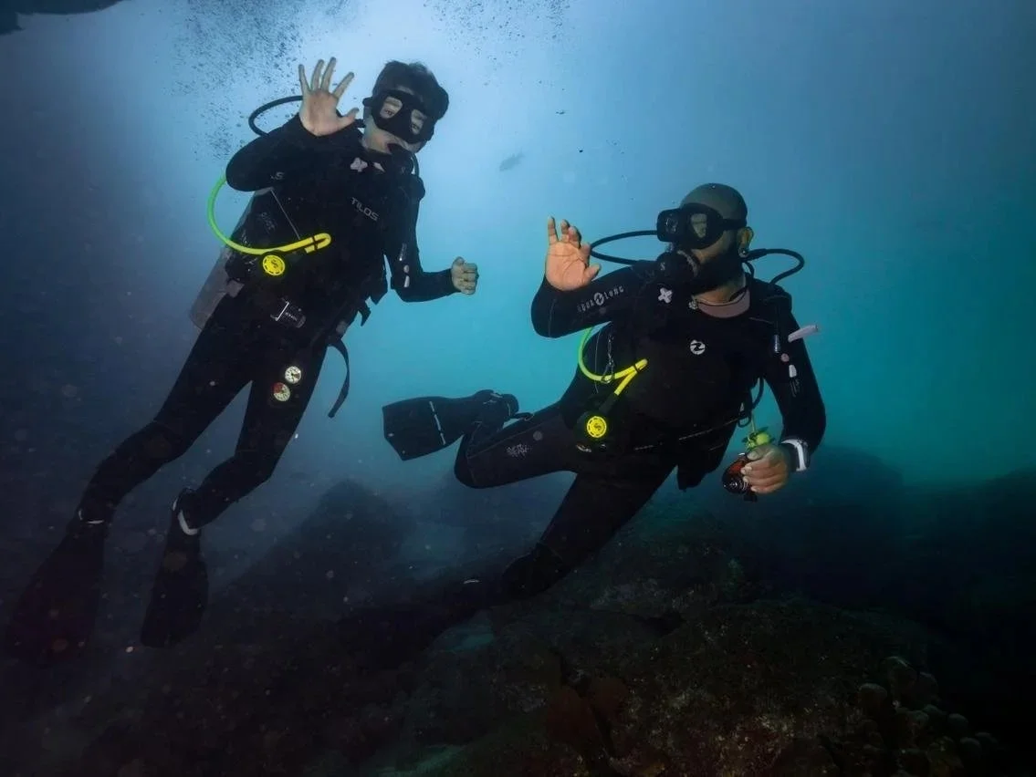 Dos buzos con equipo de buceo, actitud de saludo, en aguas profundas, alrededor de rocas.