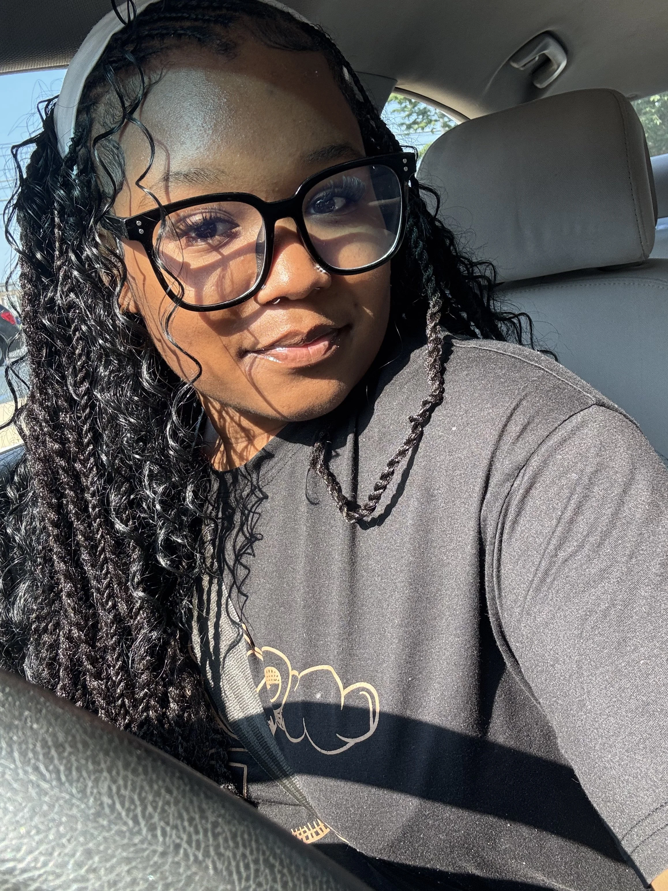 A woman with long, curly black hair, wearing large black glasses, hoop earrings, and a black T-shirt, sitting in a car and taking a selfie in sunlight.