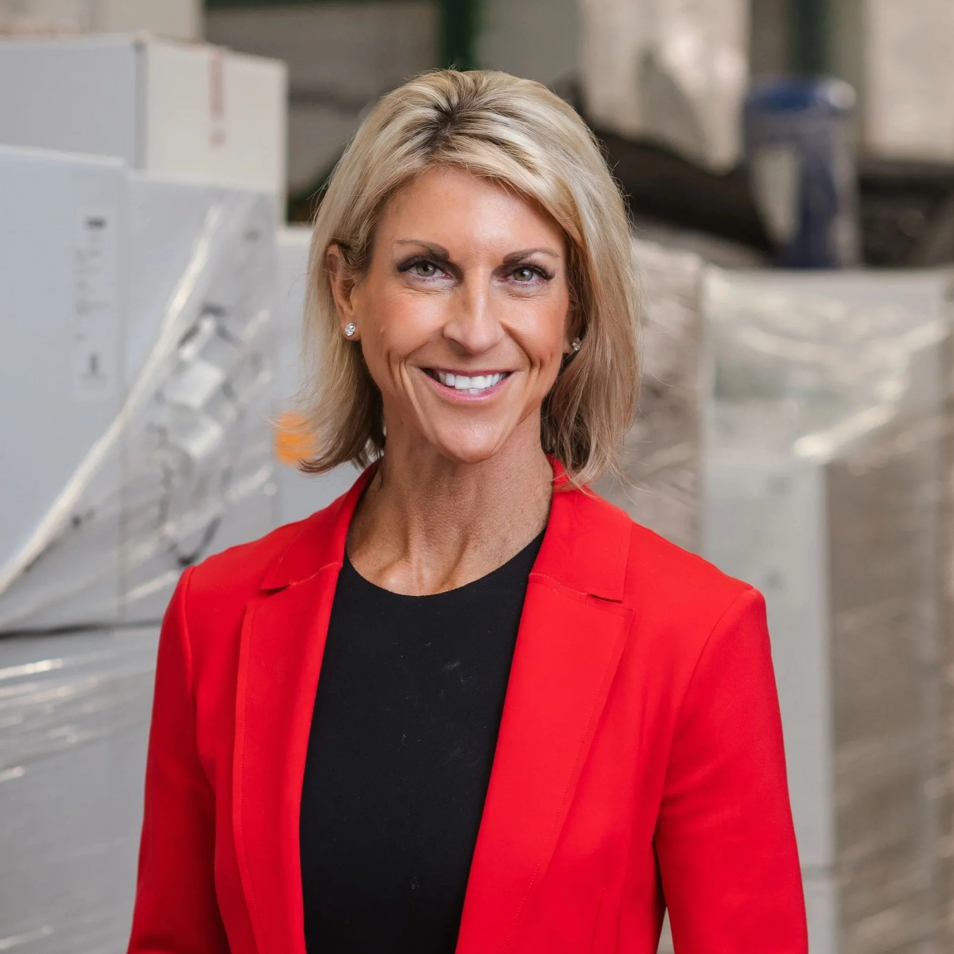 A woman with shoulder-length blonde hair, wearing a red blazer and a black top, smiling in an industrial or warehouse setting.
