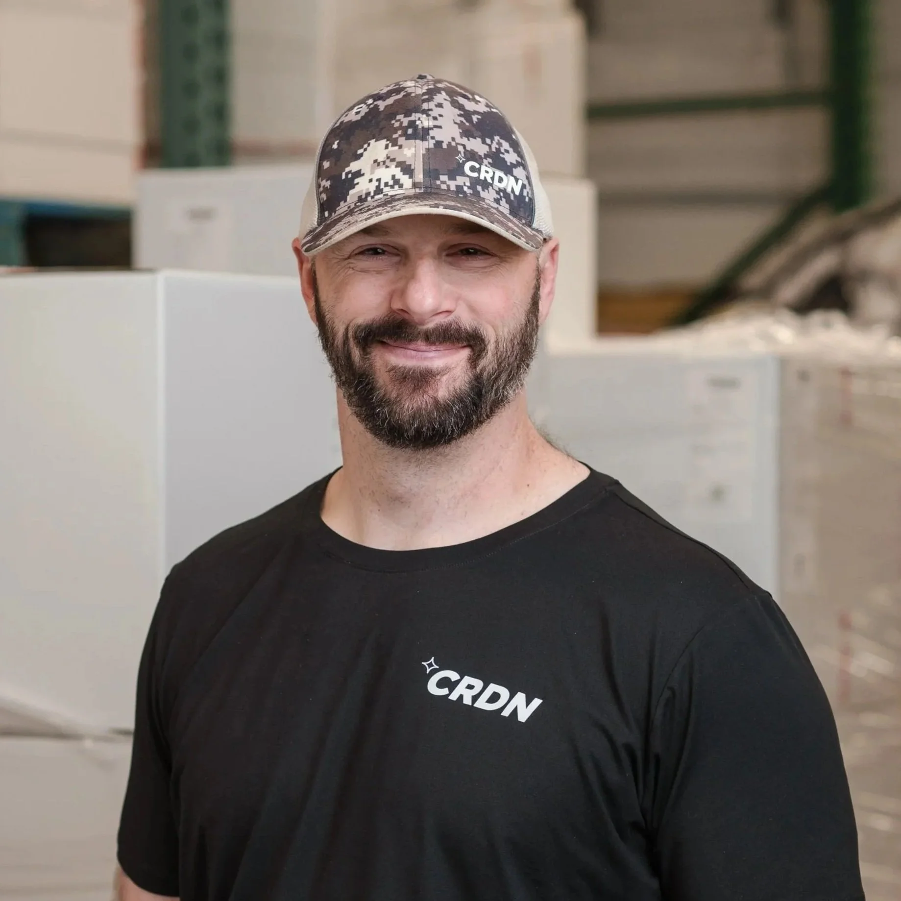 Smiling man with a beard wearing a camouflage patterned cap and a black T-shirt with 'CRDN' logo standing in a warehouse.