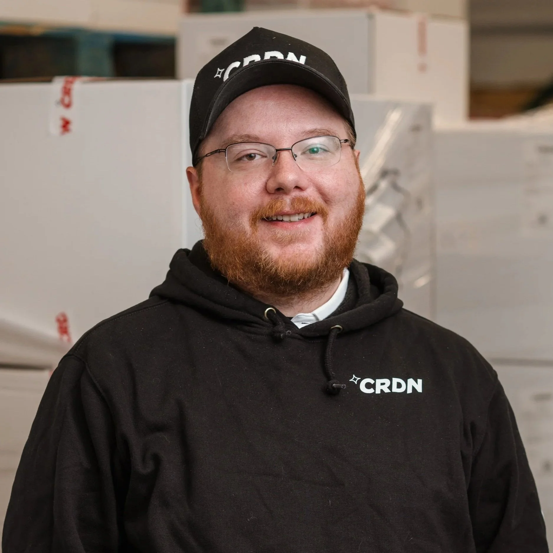 A young man with glasses, a red beard, and a black hoodie with 'CRDN' logo, smiling in an industrial or warehouse setting with cardboard boxes in the background.