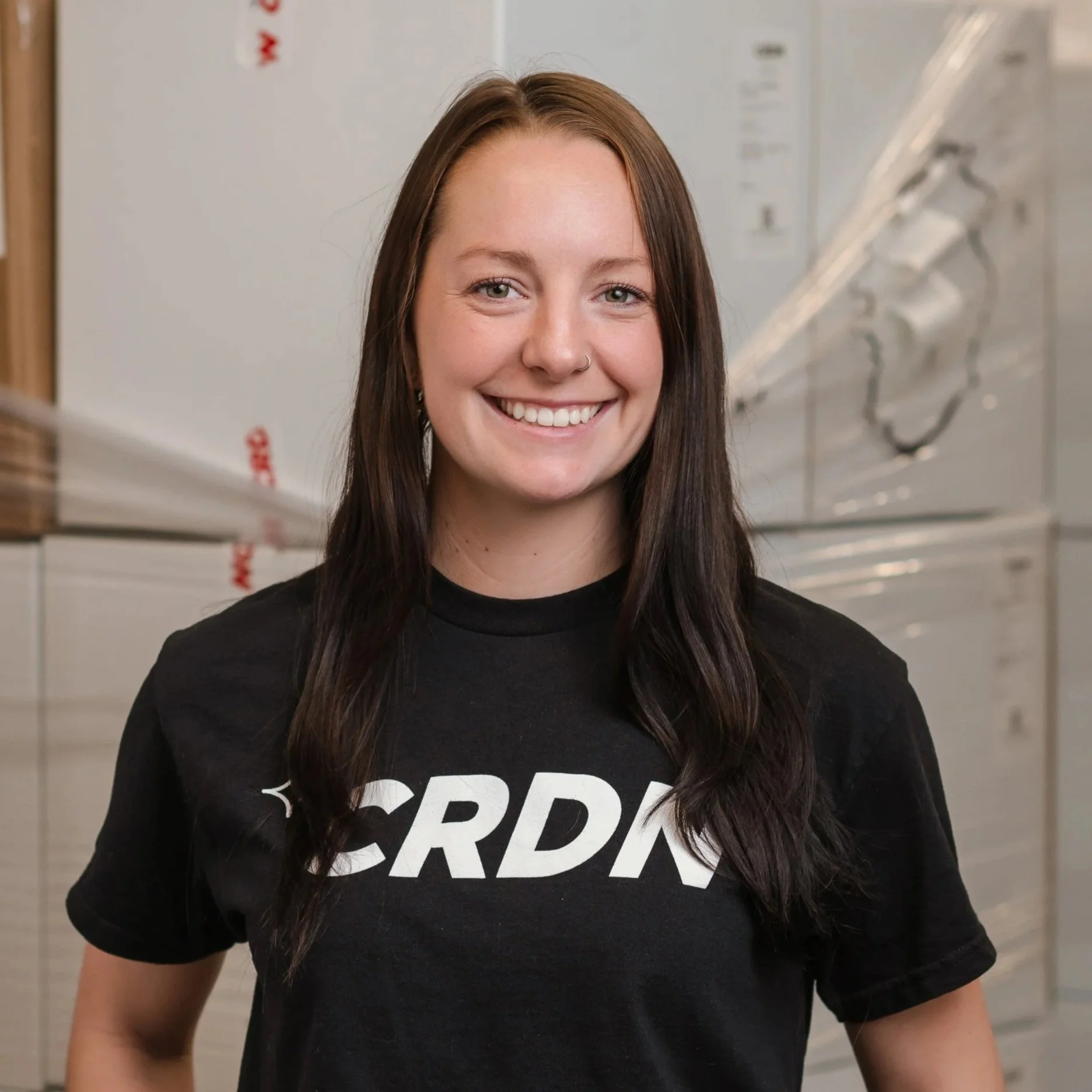 A young woman with long dark hair, smiling, wearing a black T-shirt with white text, standing indoors in front of stacked boxes.