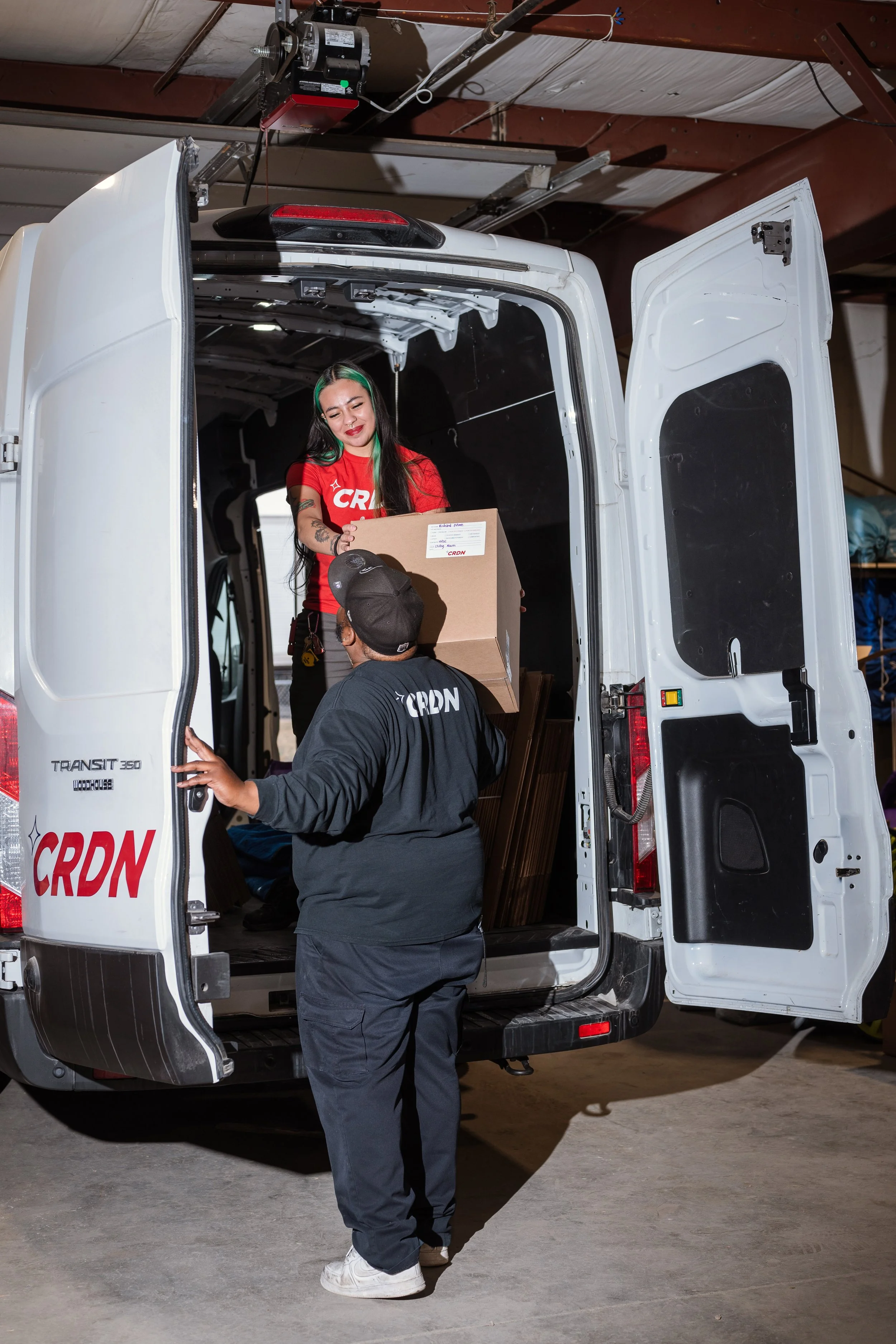 Two delivery workers loading boxes into the back of a white delivery van. The woman is on the van's step handing a box to the man, both wearing Black and Red uniforms with the logo 'CRDN.' The van is parked inside a warehouse or garage.