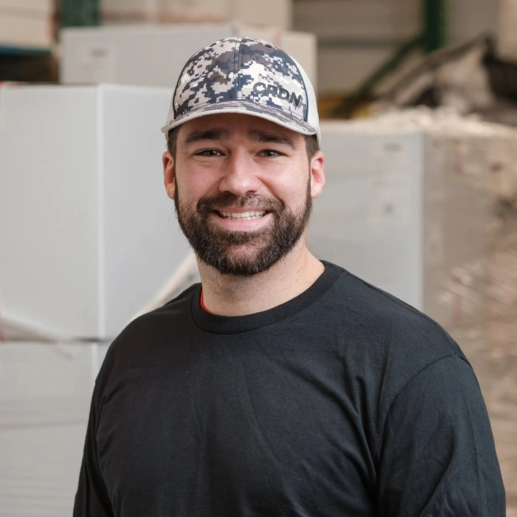 A smiling man with a beard wearing a camouflage baseball cap and a black shirt, standing in an indoor industrial or warehouse setting