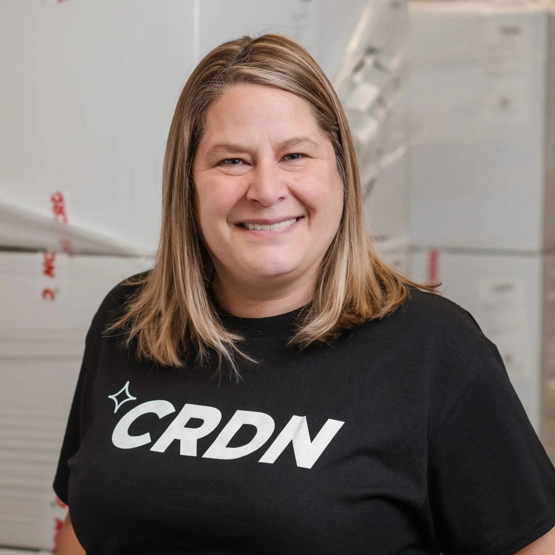 A smiling woman with shoulder-length light brown hair, wearing a black t-shirt with the word 'CRDN' printed on it, standing in front of boxes in an industrial setting.