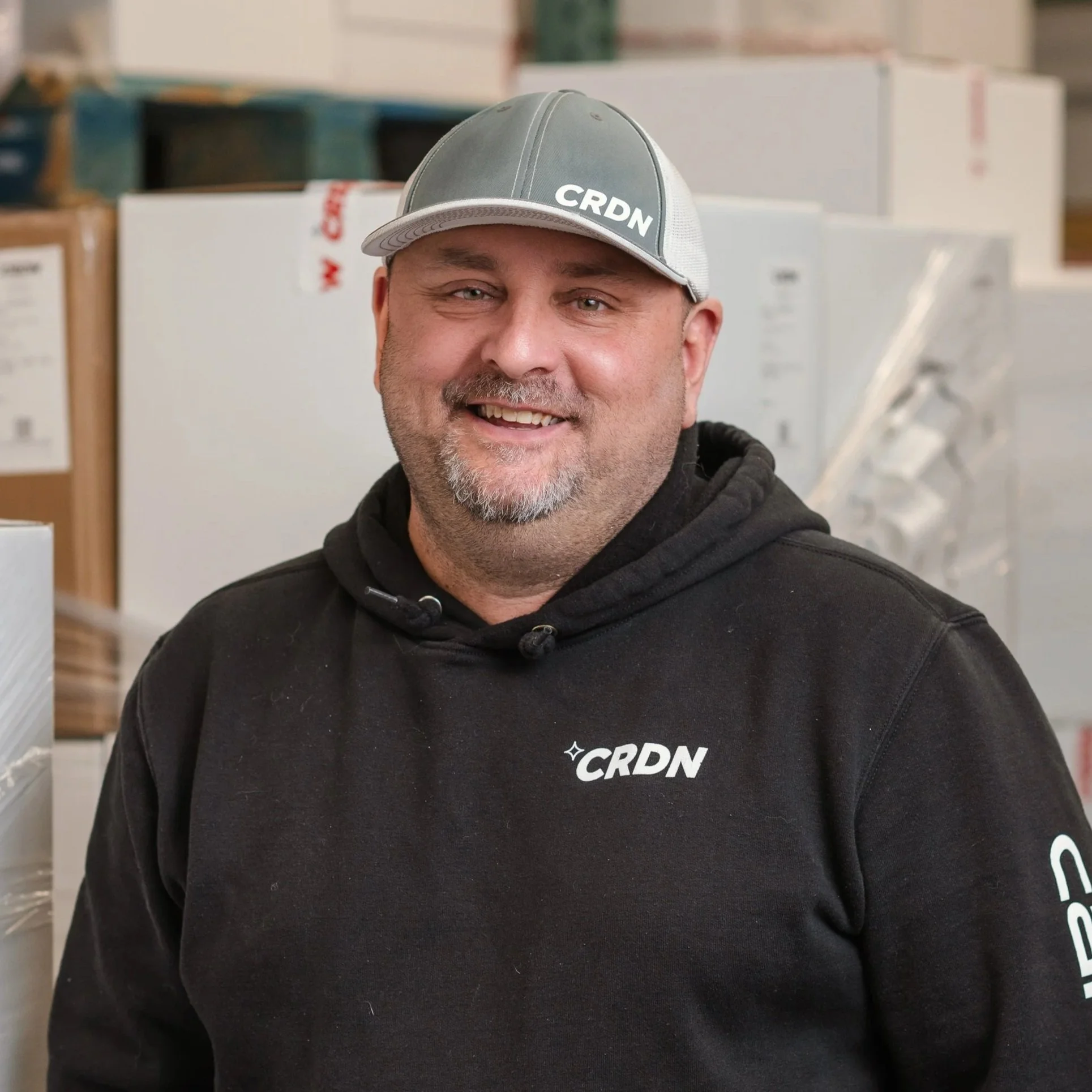 A smiling man with a beard and mustache wearing a gray and black baseball cap and black hoodie with white 'CRDN' logos, standing in a warehouse with boxes and shelves in the background.