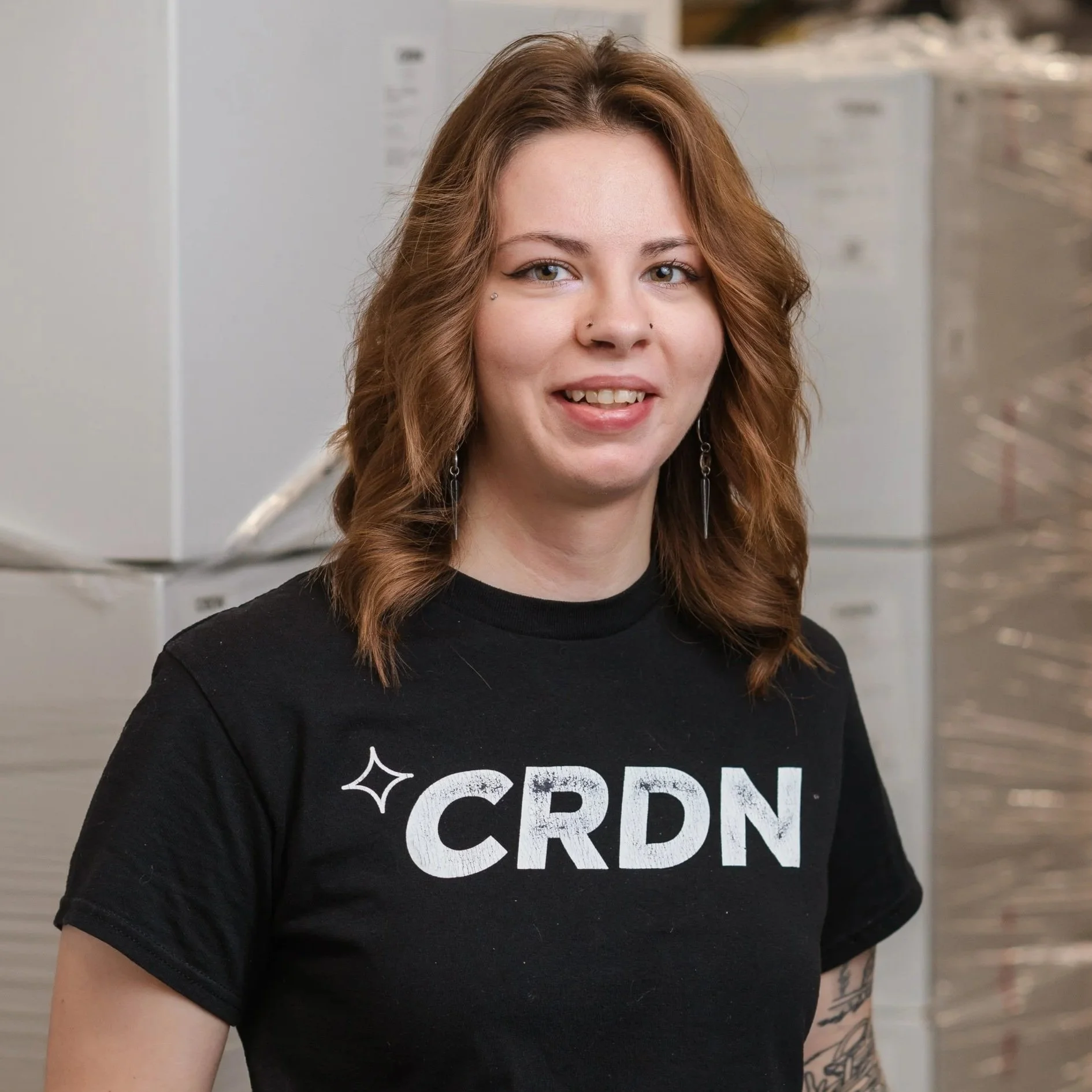 A young woman with shoulder-length wavy red hair, wearing a black t-shirt with 'CRDN' printed on it, standing in an industrial setting with metallic stacked storage boxes in the background.