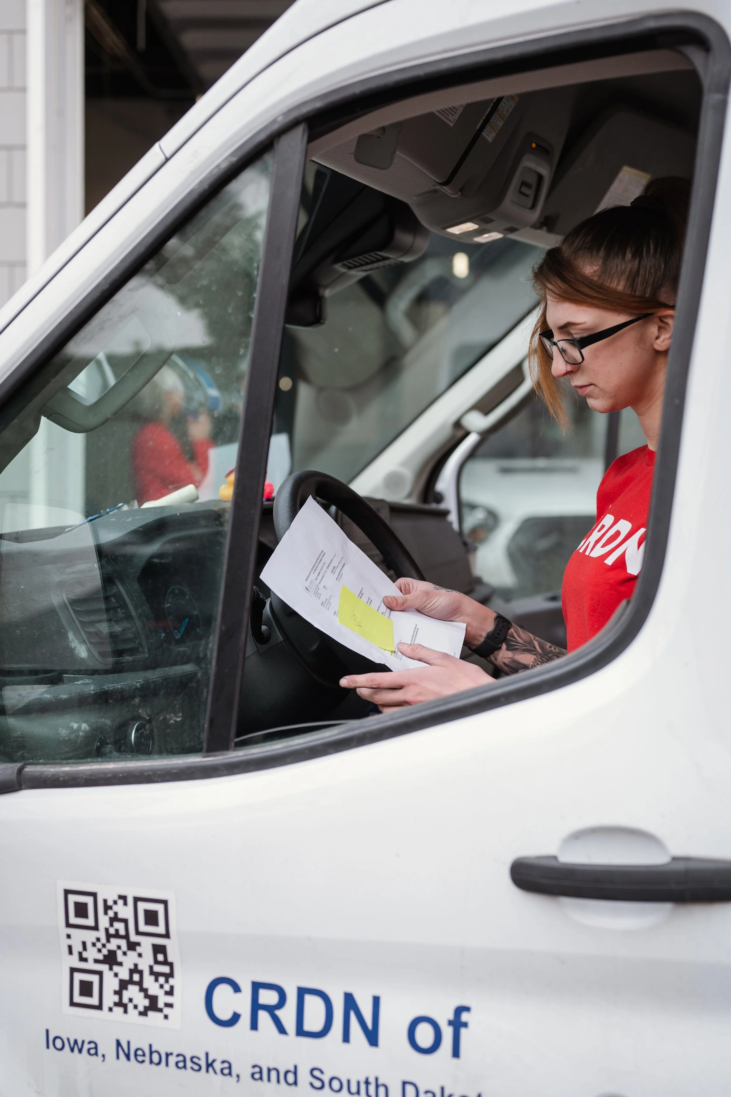 A woman with glasses and tattoos is sitting in the driver's seat of a white delivery van, reading documents. The van has a QR code and text on the side indicating it is part of CRDN of Iowa, Nebraska, and South Dakota.