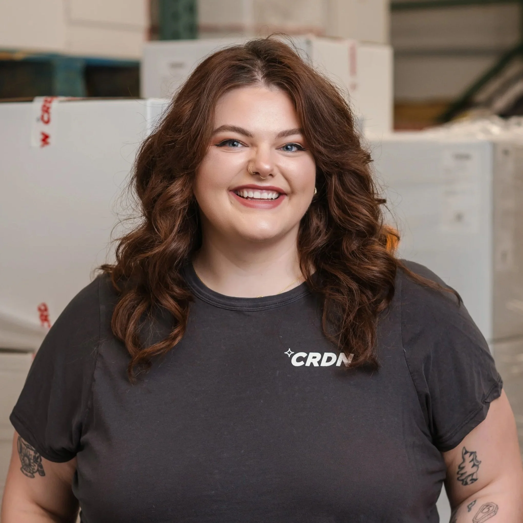A young woman with red hair, smiling, wearing a black t-shirt with 'CRDN' logo, standing in front of stacked boxes.
