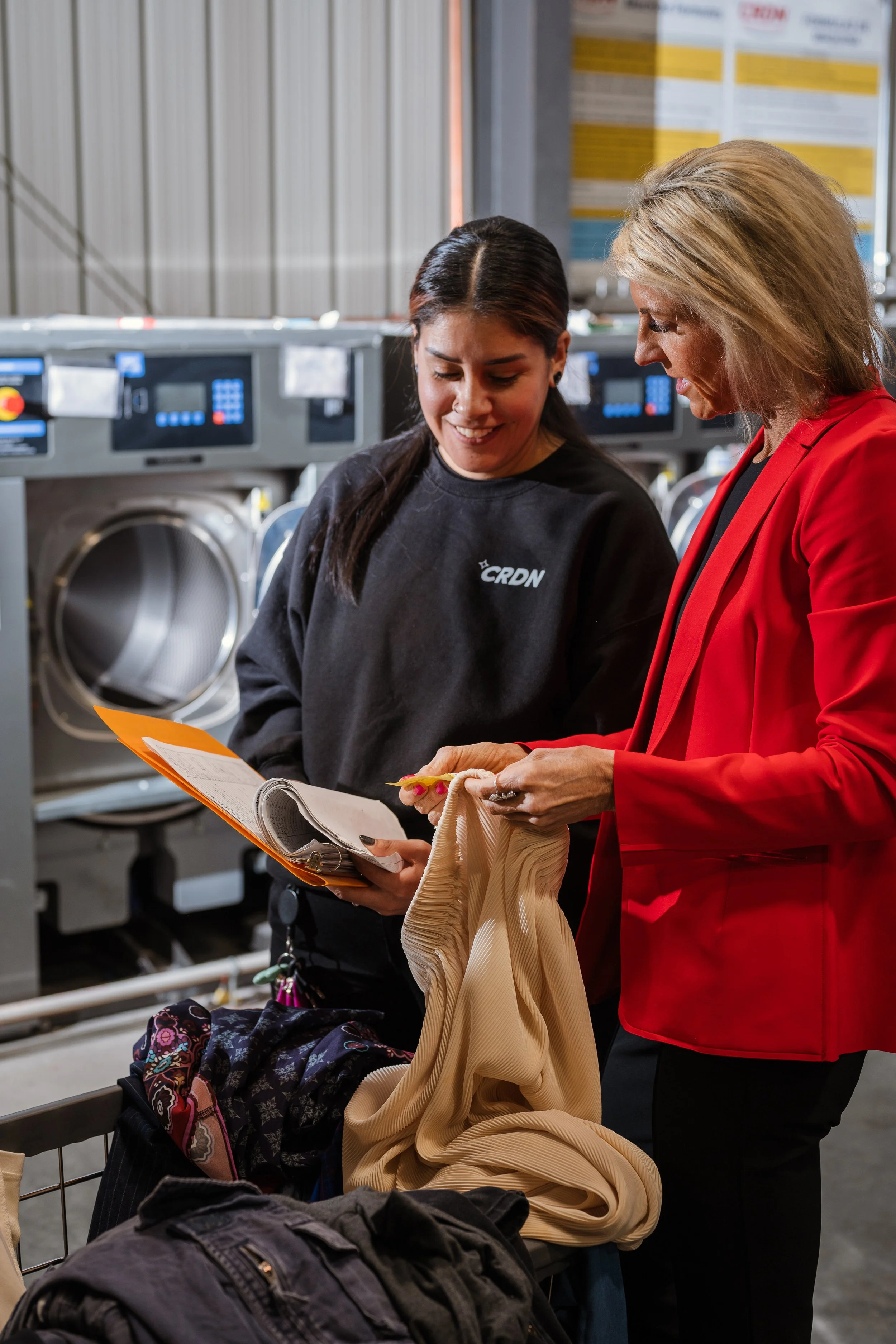 Two women in a coin laundry, discussing while looking at a clipboard and a clothing fabric.