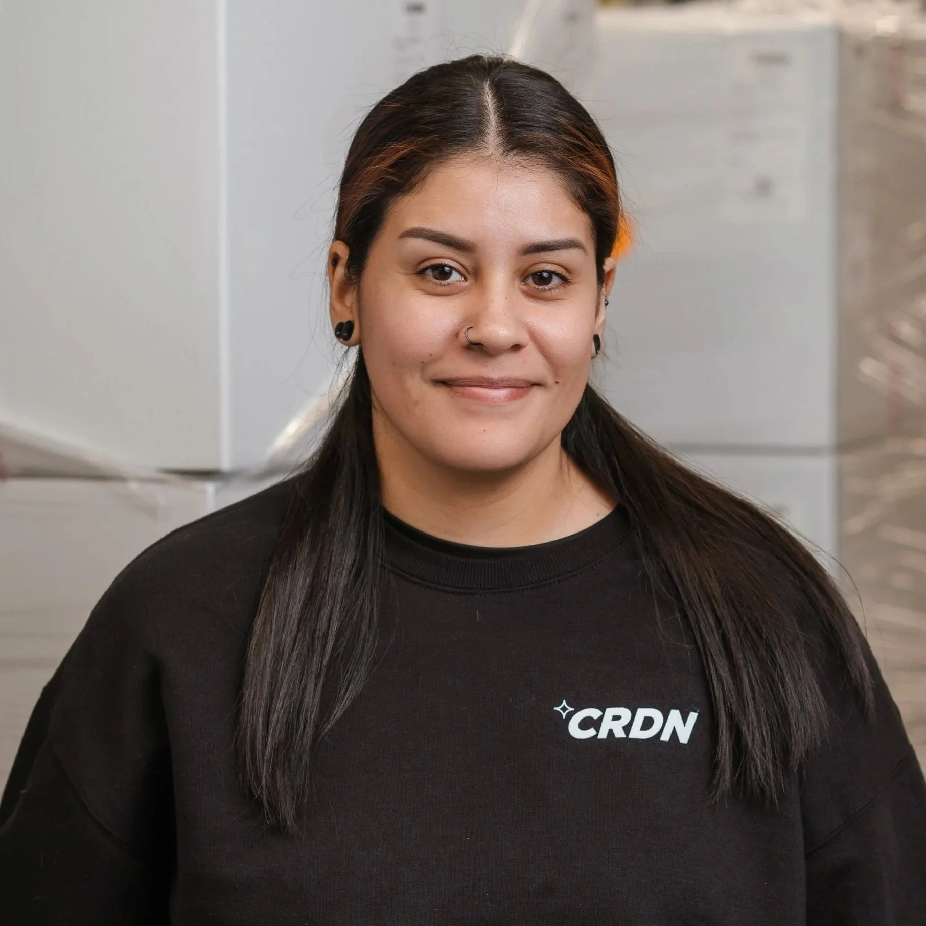A young woman with dark hair and a nose ring, wearing a black sweatshirt with 'CRDN' logo, standing in front of stacked boxes.