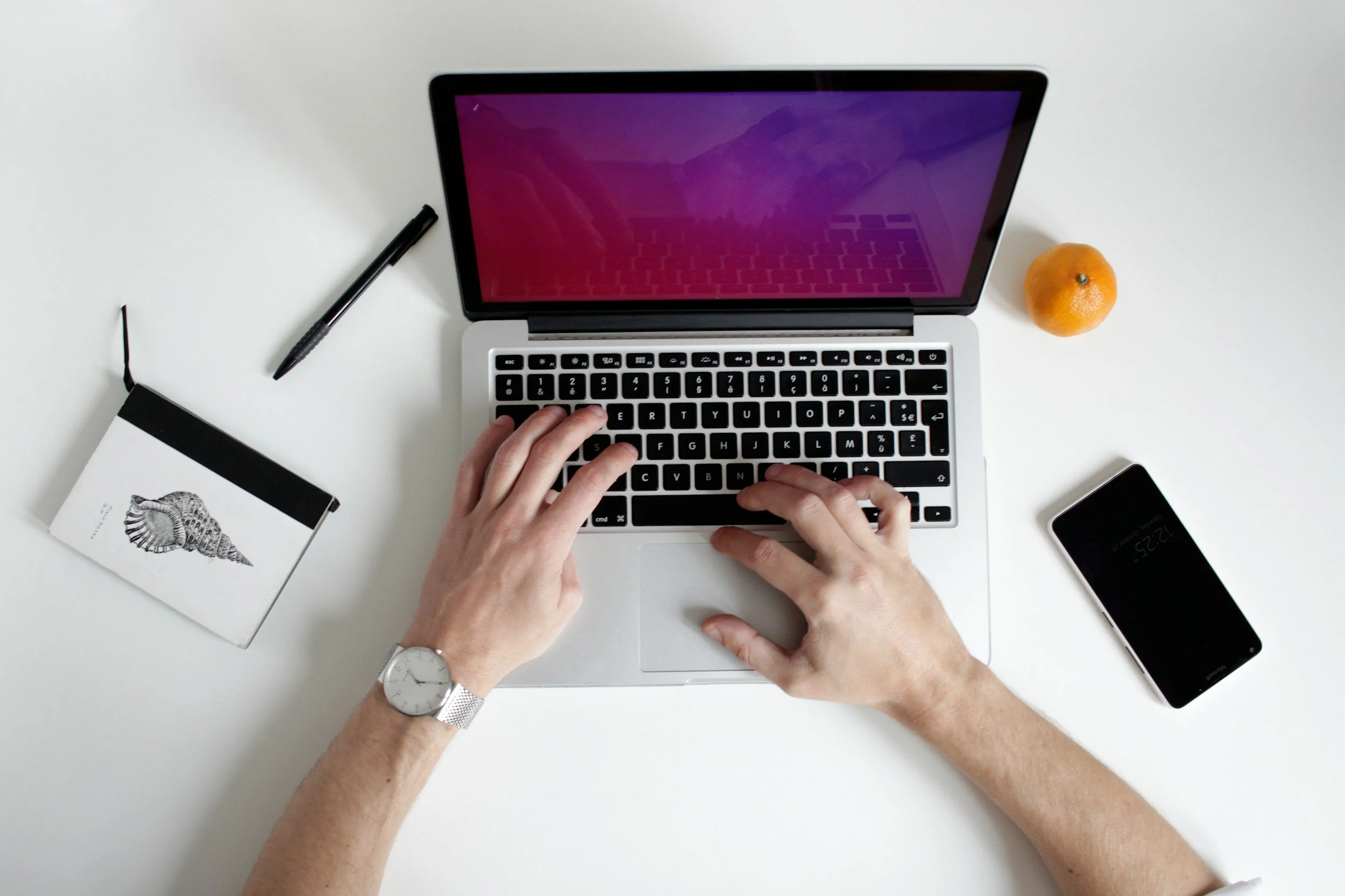 Top view of a person typing on a silver laptop with a pink and purple screen on a white desk. The person is wearing a silver watch on their left wrist. Nearby, there is a black pen, a small notebook with a seashell drawing, an orange, and a black smartphone.