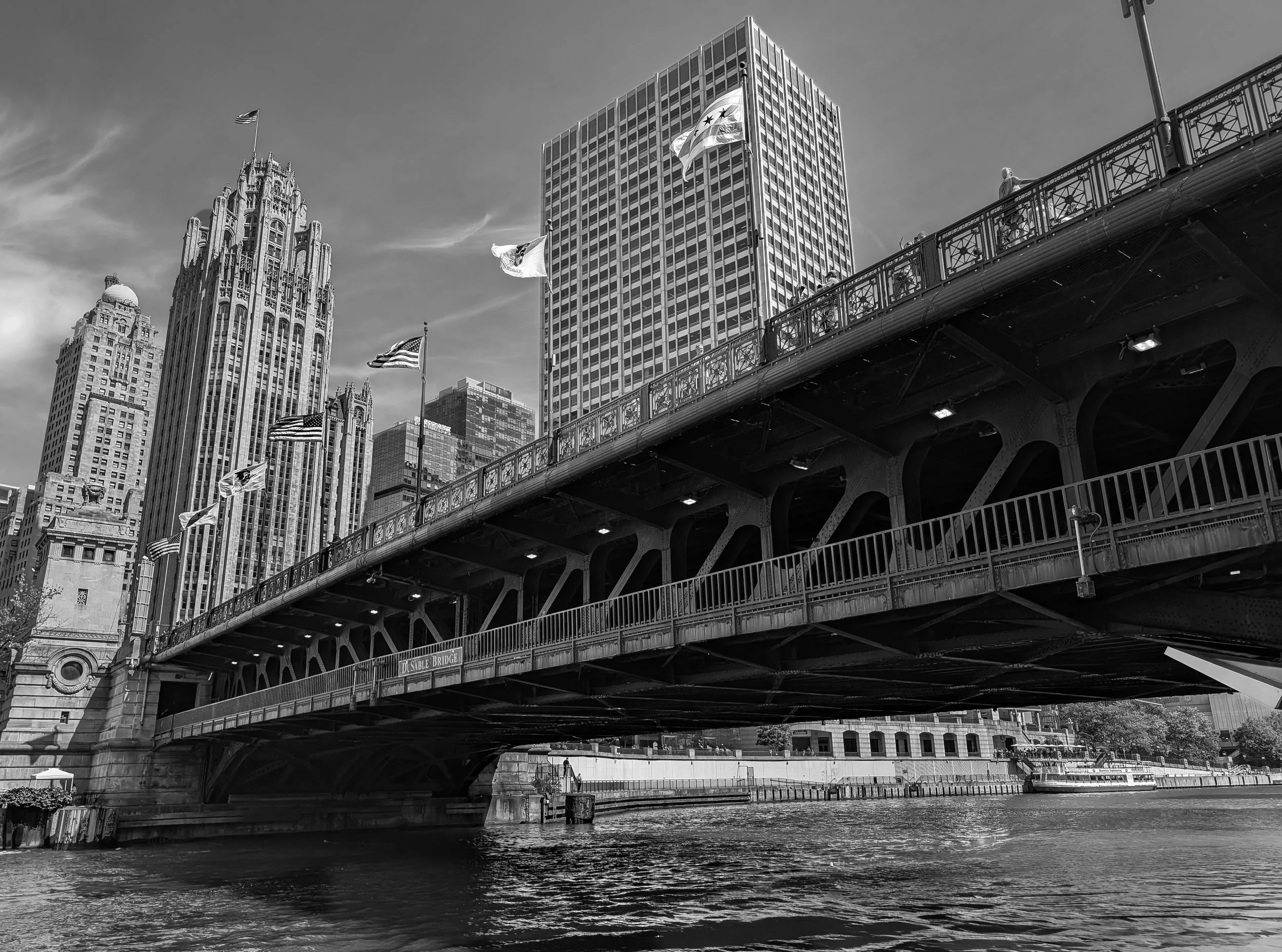 Black and white photo of city skyline with tall buildings, a bridge over a river, and flags flying on the bridge.