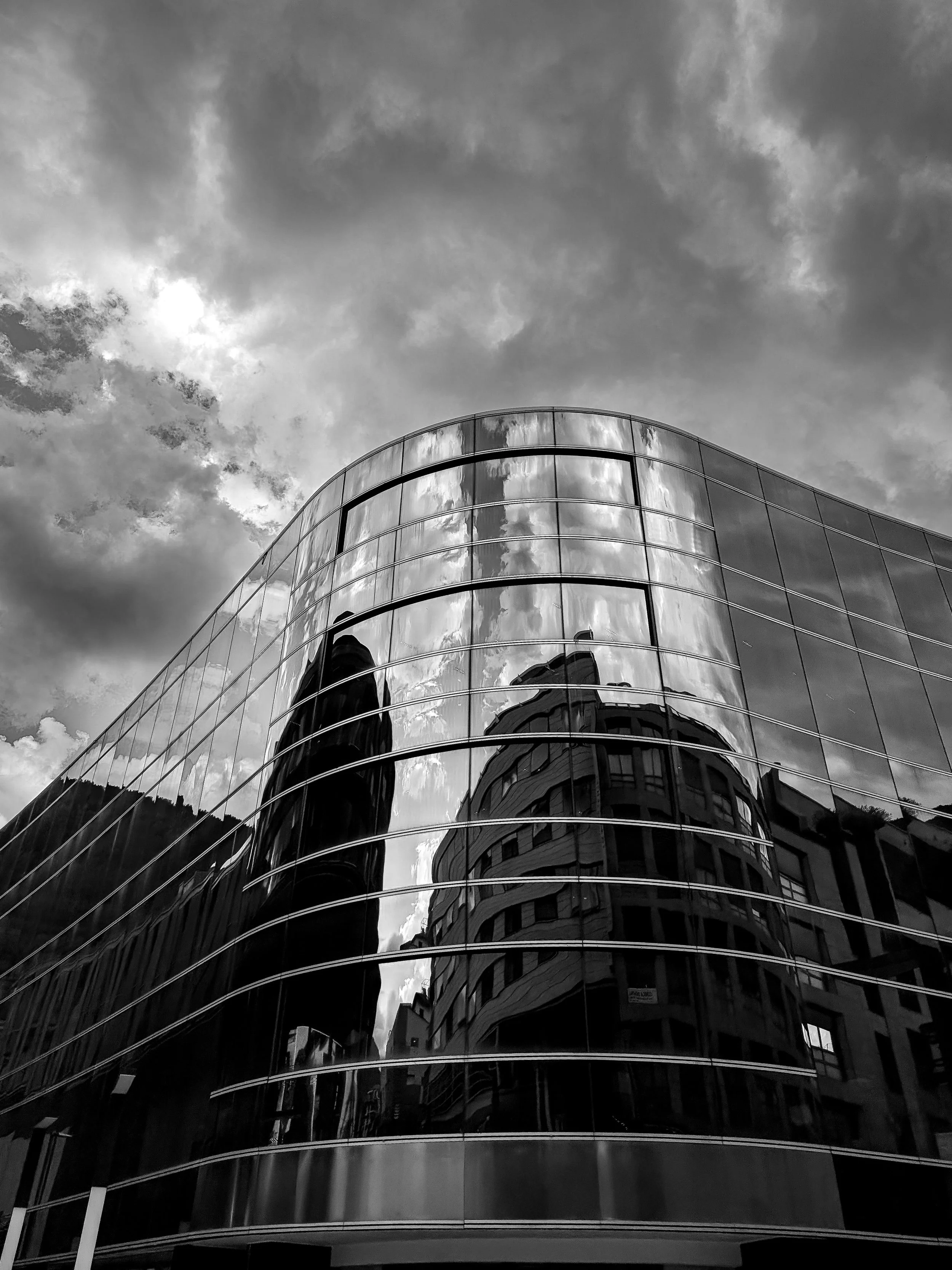 Black and white photo of a modern glass building reflecting the cloudy sky and nearby structures.